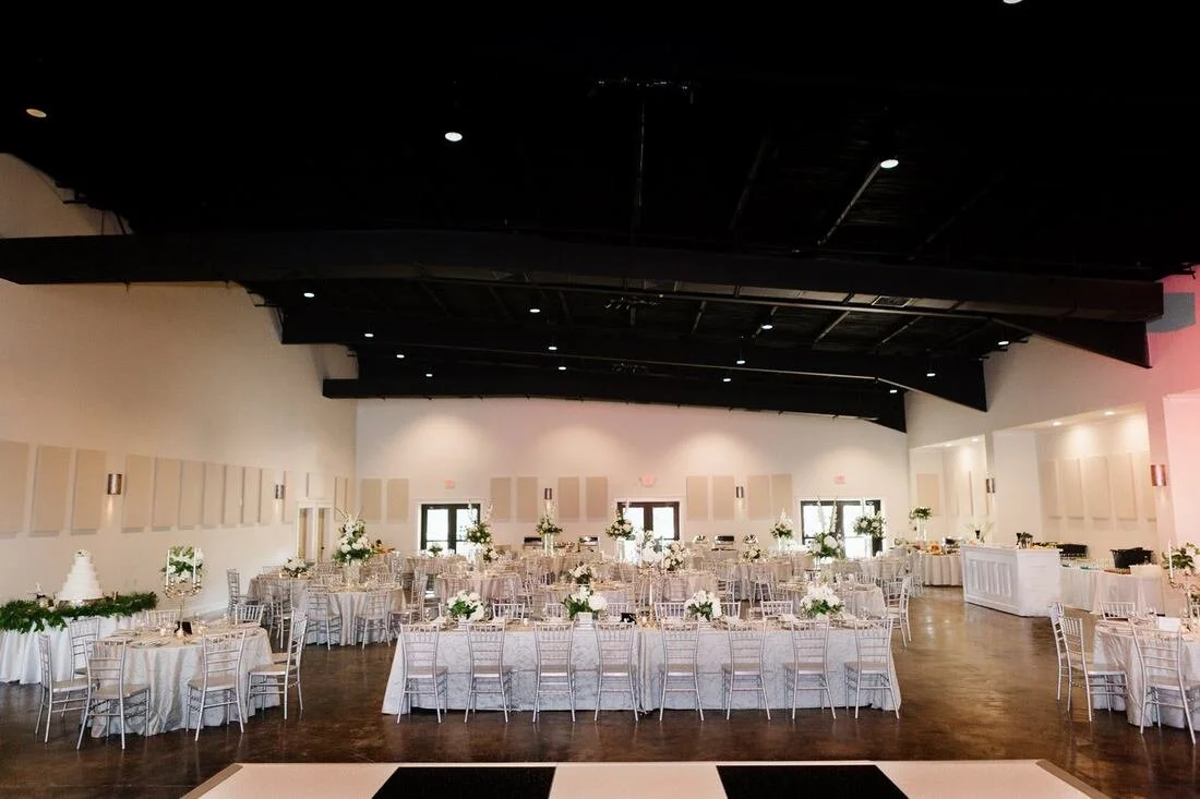 Wedding reception hall with arranged tables and chairs, floral centerpieces, a multi-tiered wedding cake on a side table, and a white bar area, prepared for guests.