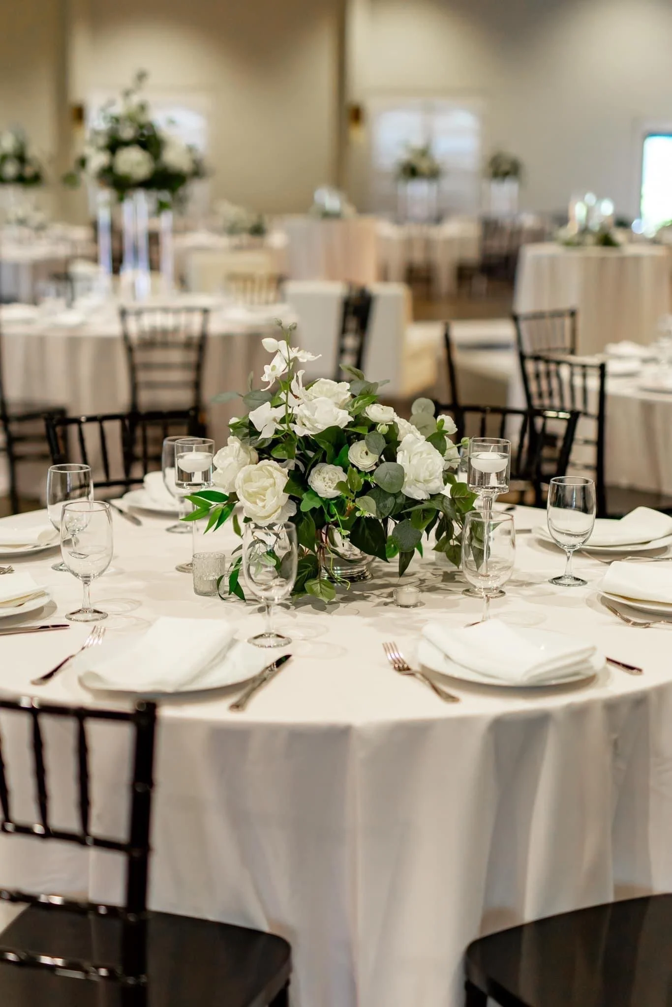 Elegant banquet table with a floral centerpiece, white tablecloth, napkins, wine glasses, water glasses, and silverware in a decorated event hall.