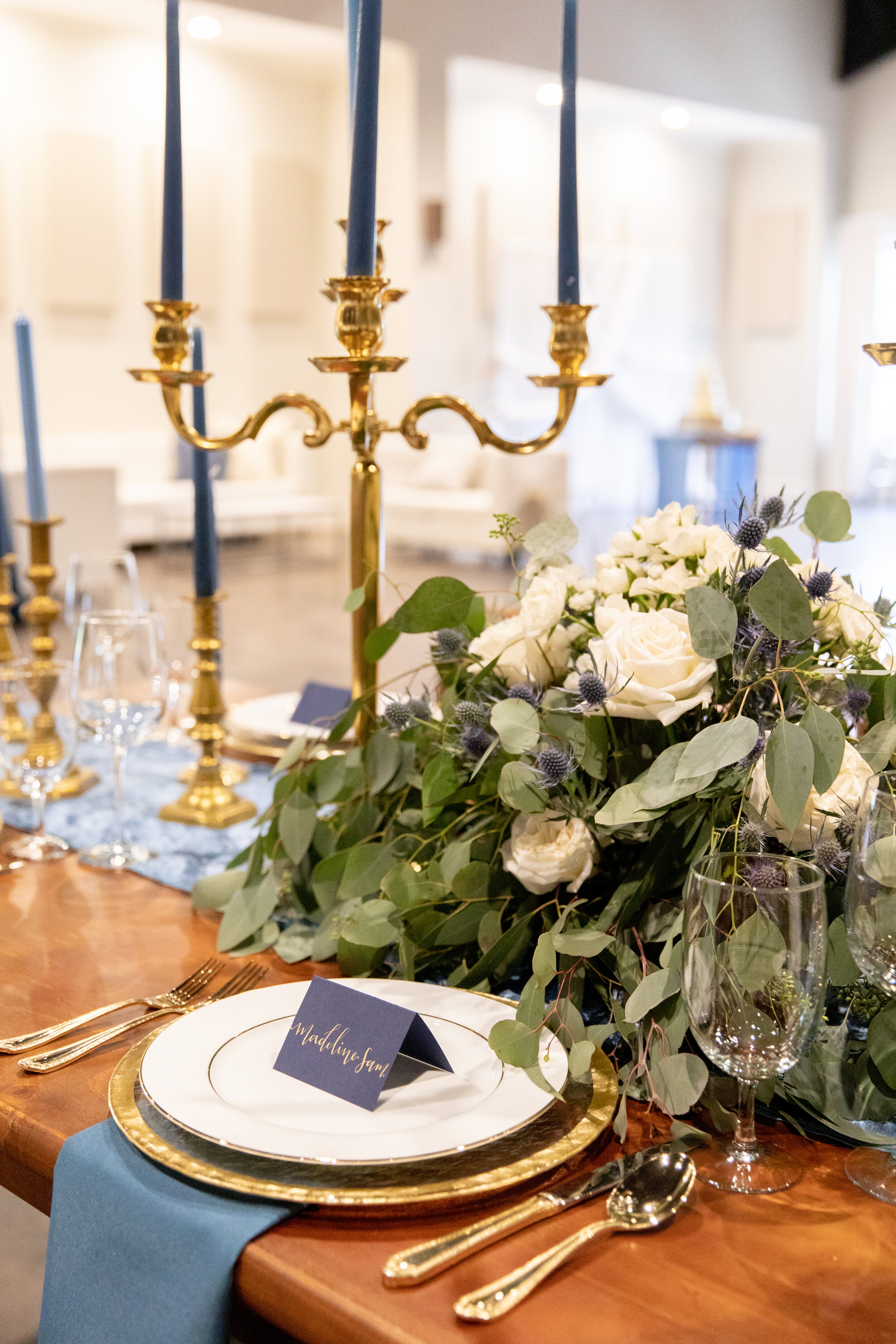 A formal dining table setting with gold-rimmed plates, gold utensils, blue napkins, and a floral centerpiece with white roses and greenery, accented by a gold candelabra with blue candles.