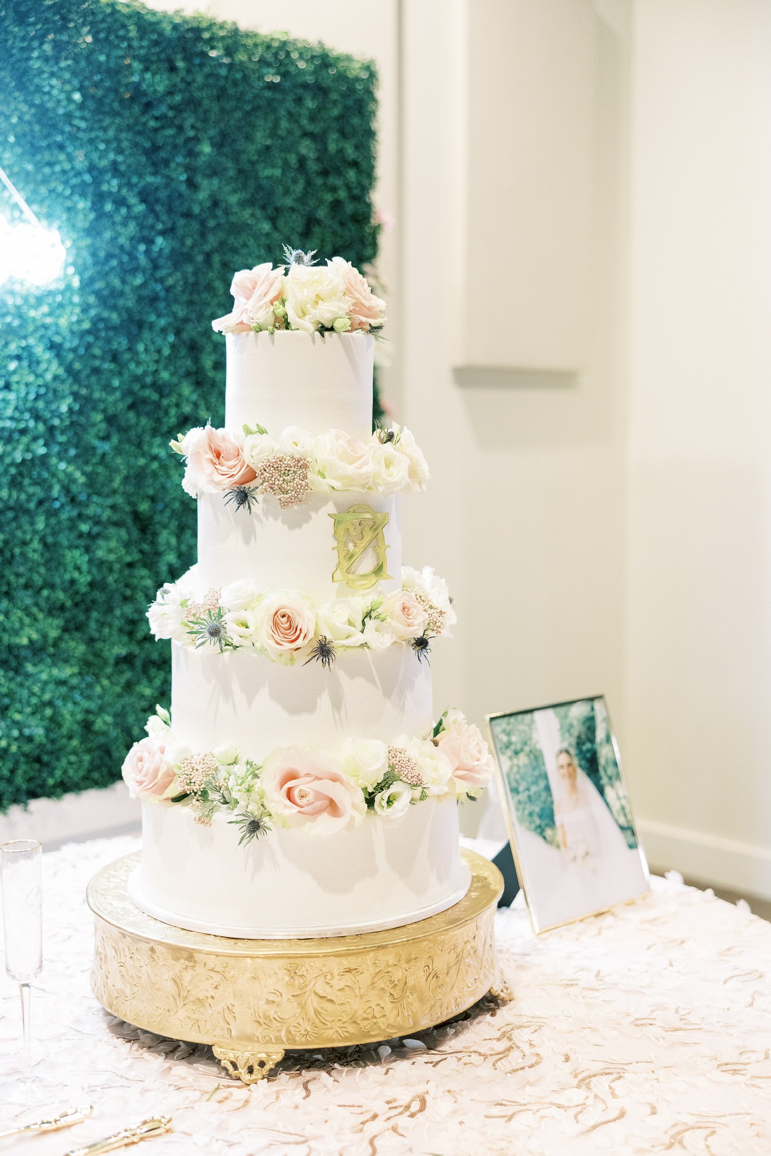 A four-tier white wedding cake decorated with pink and white flowers, displayed on an ornate gold cake stand, with a framed photograph of a bride and a green leafy backdrop in the background.