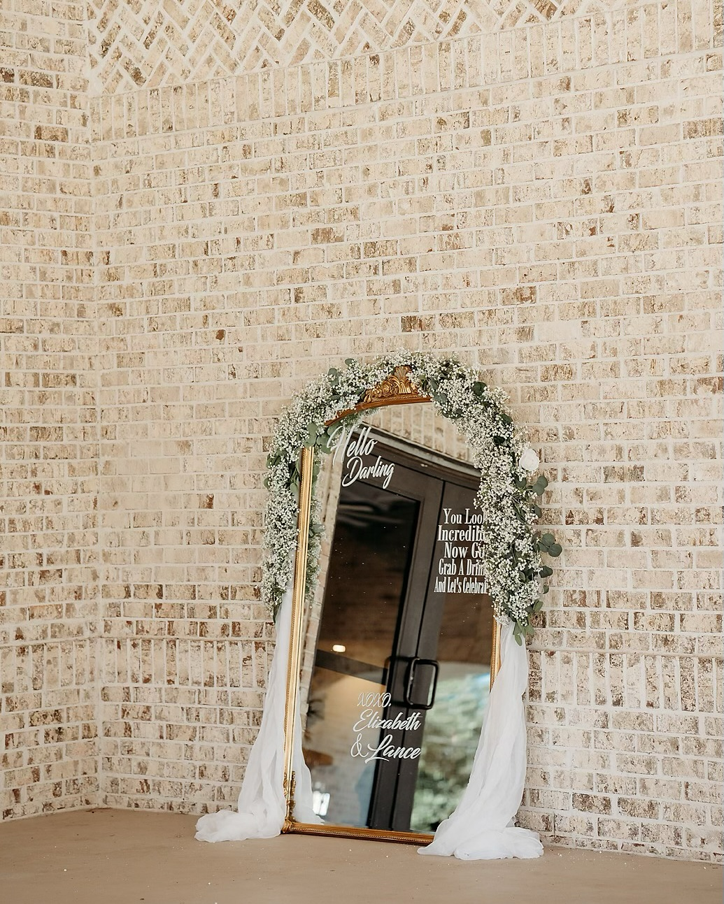 Decorative wedding mirror with white floral garland and white fabric draping against a brick wall, with white scripted text on the mirror reading 'Hello Darling' and a message about a celebration for Elizabeth and Lance.