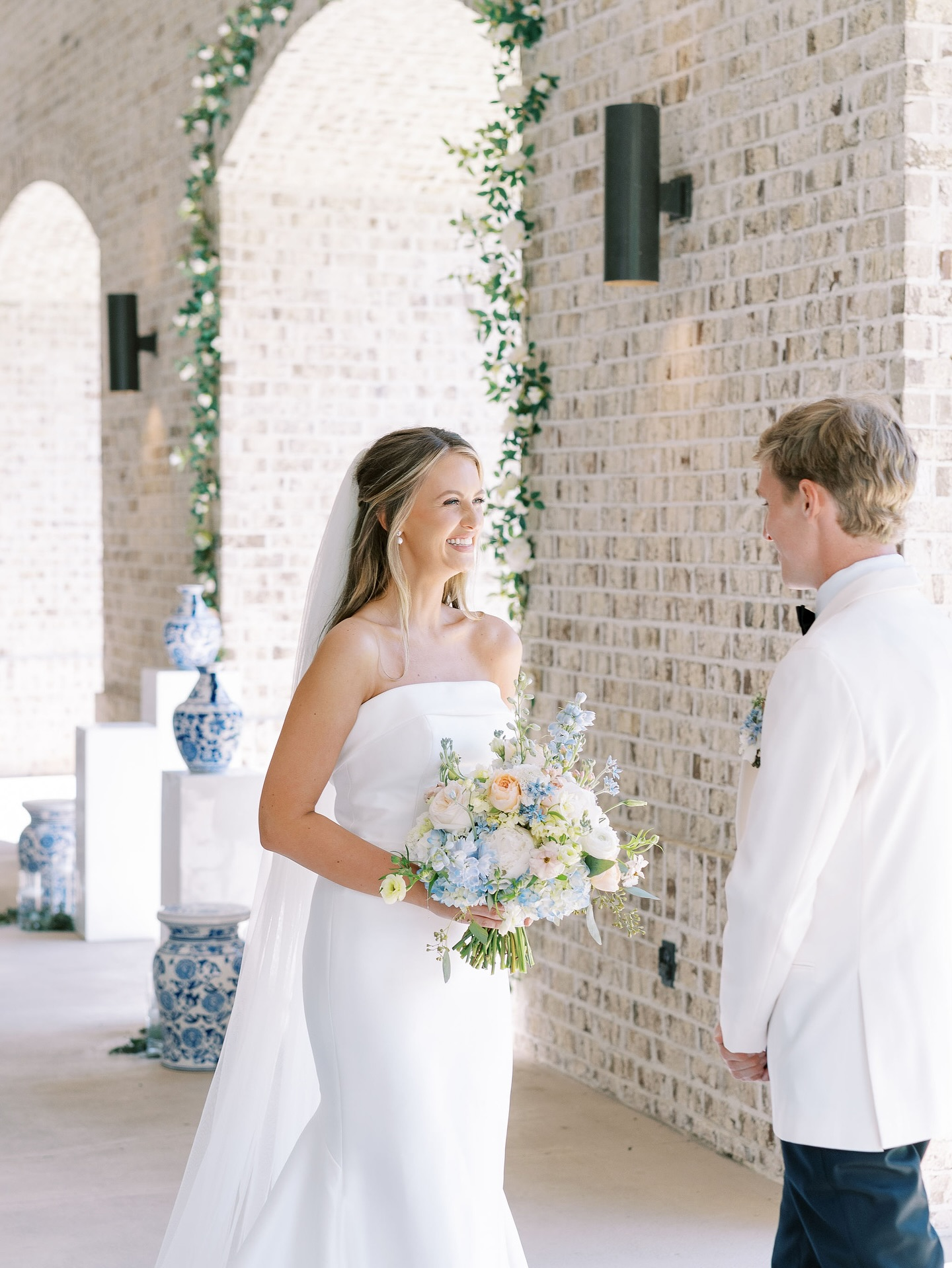 A bride and groom on their wedding day standing indoors by a brick wall, with the bride holding a bouquet of pastel flowers and smiling at the groom.