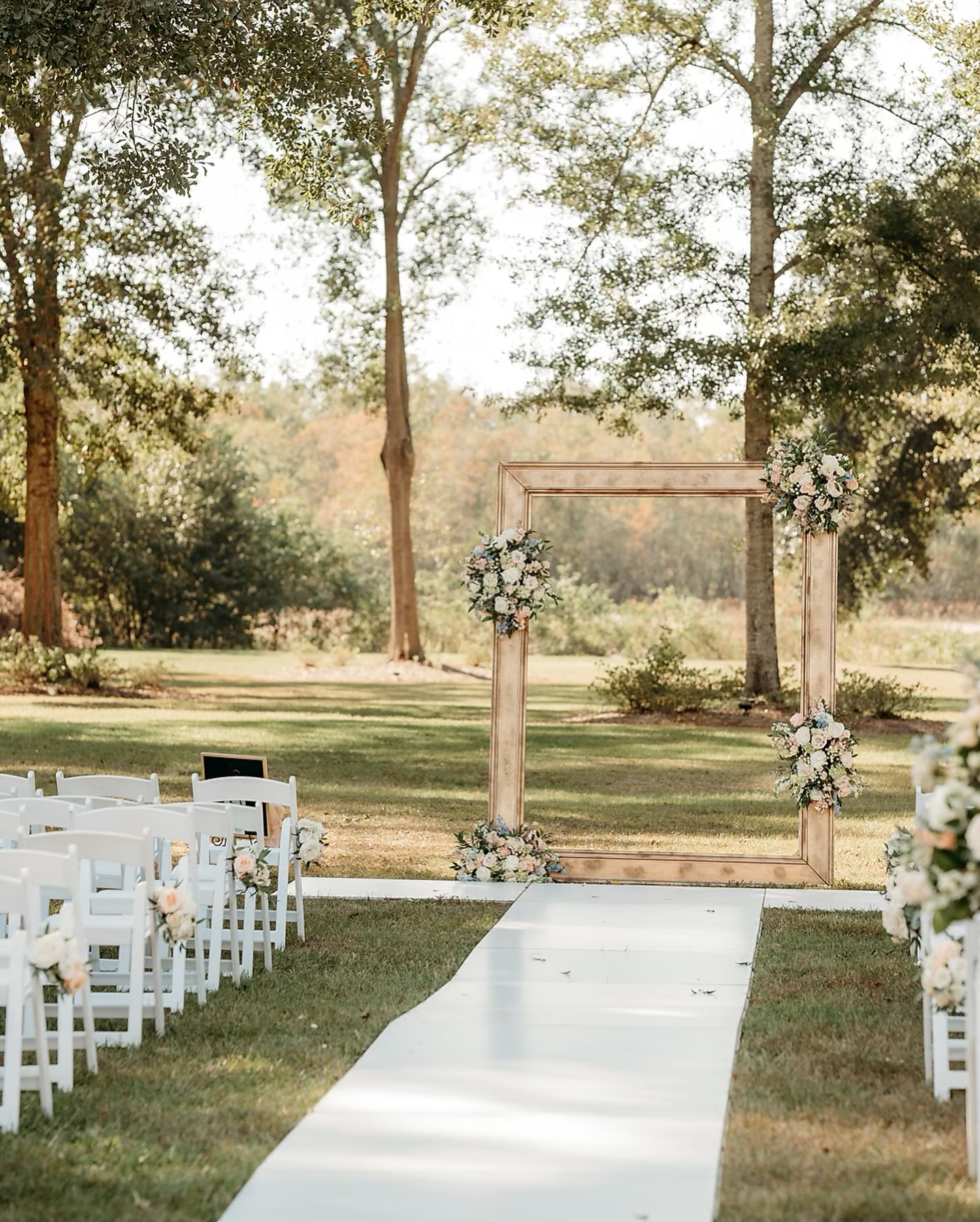 Outdoor wedding ceremony setup with white chairs decorated with floral arrangements, a white aisle runner, and a wooden frame decorated with flowers for an altar, set in a sunny, green park with trees in the background.