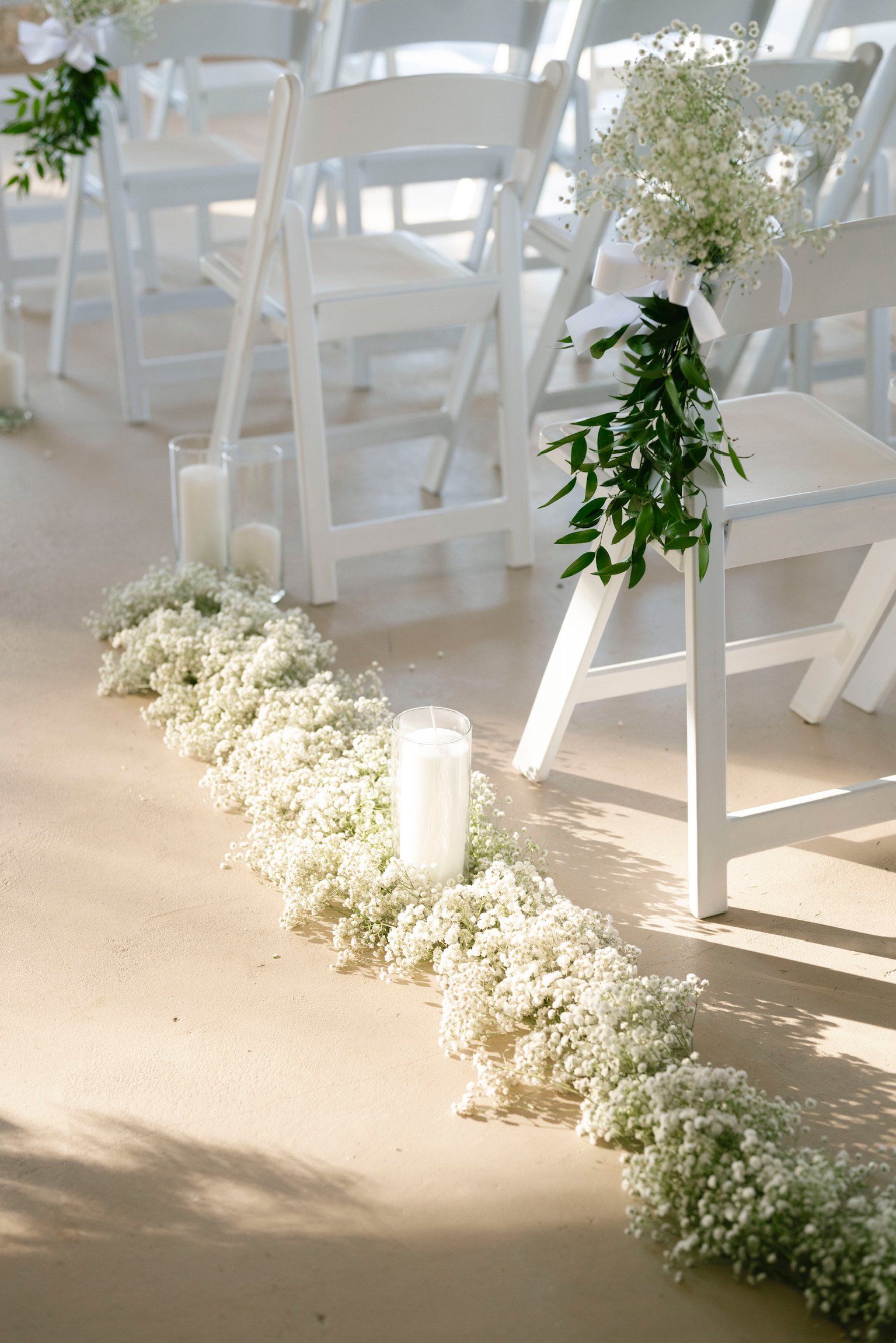 White folding chairs decorated with greenery and white flowers, lining a sandy aisle with white floral arrangements and candles for a wedding ceremony.