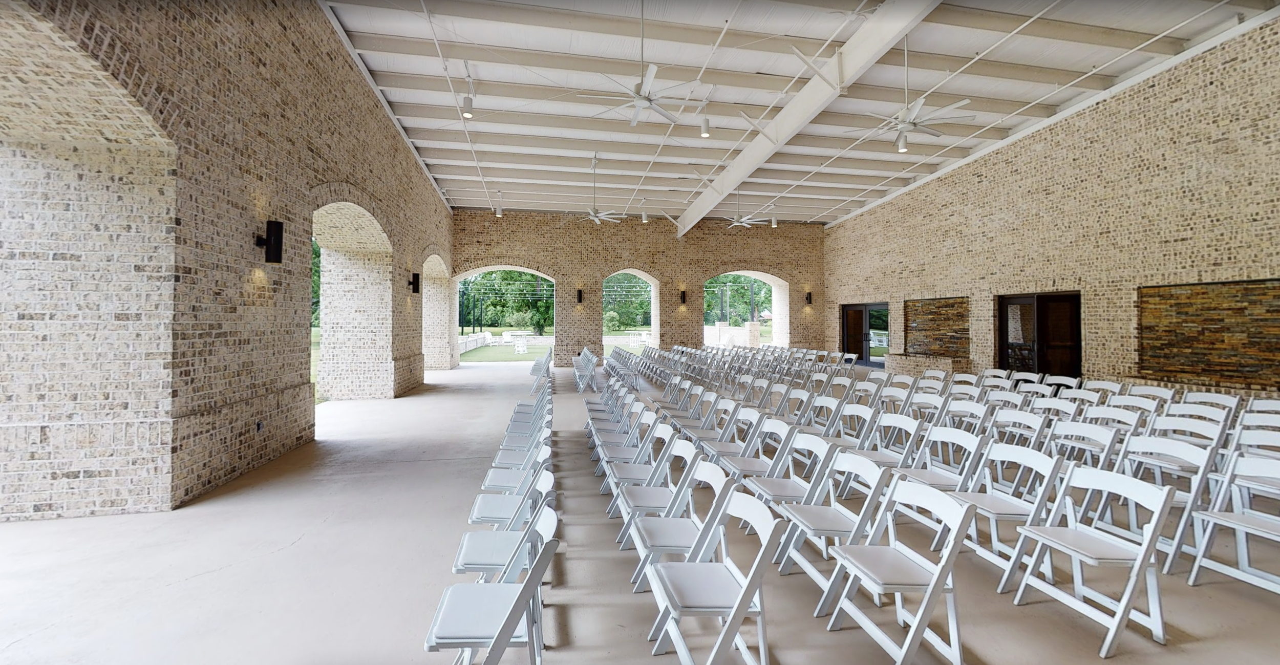 Indoor event space with rows of white chairs arranged facing forward, brick walls, large arched windows with view of greenery outside, ceiling fans, and wall-mounted lights.