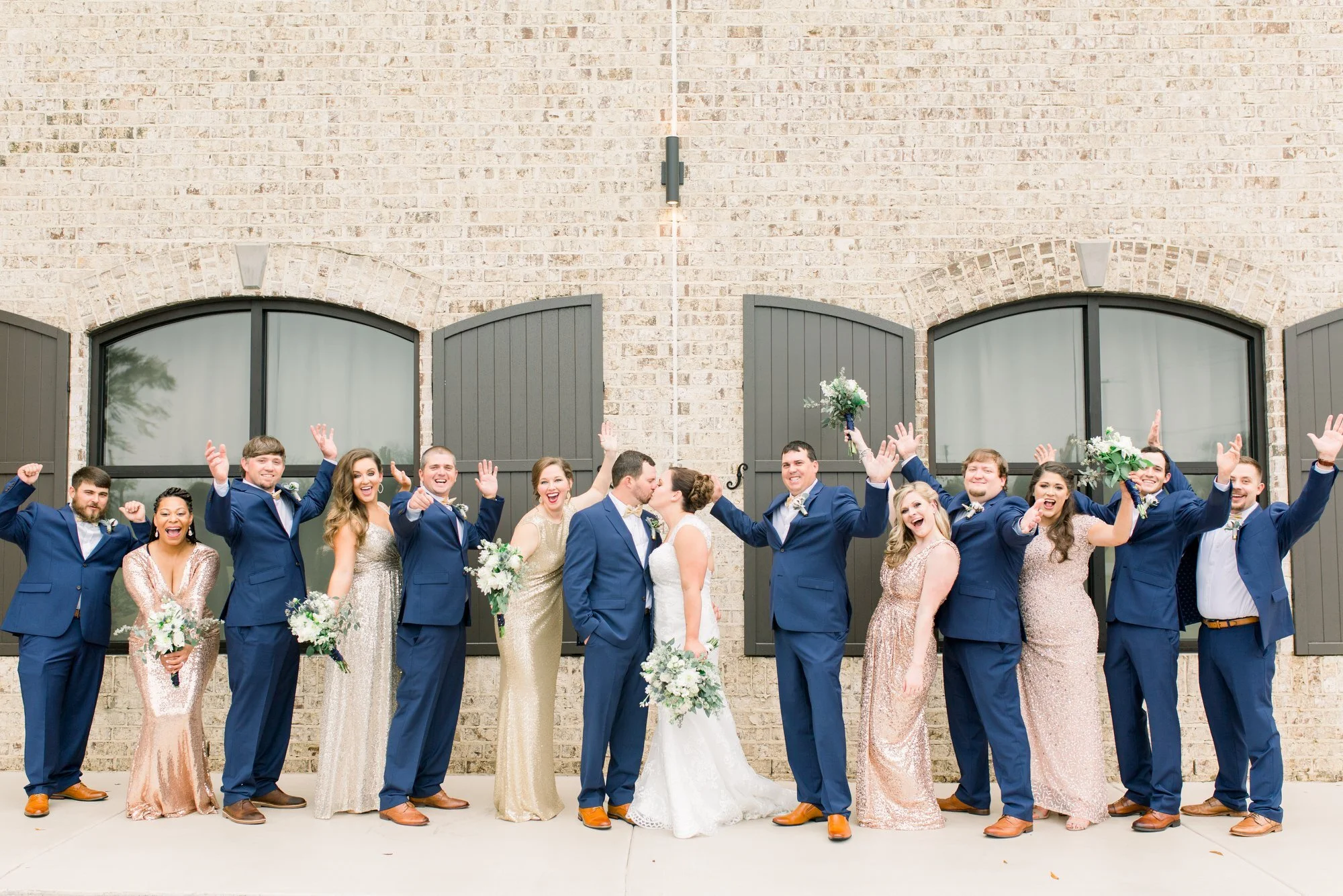 A group of people in wedding attire celebrating outdoors, with a couple kissing in the center, surrounded by friends holding bouquets and raising their hands, against a brick wall with large windows and dark green shutters.