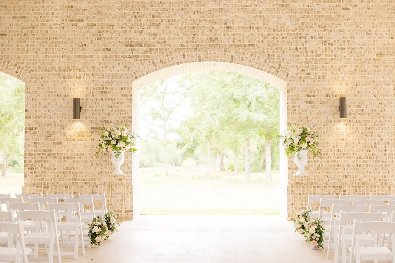Wedding ceremony setup inside a brick chapel with white chairs and floral arrangements on either side, leading to an outdoor view of trees.
