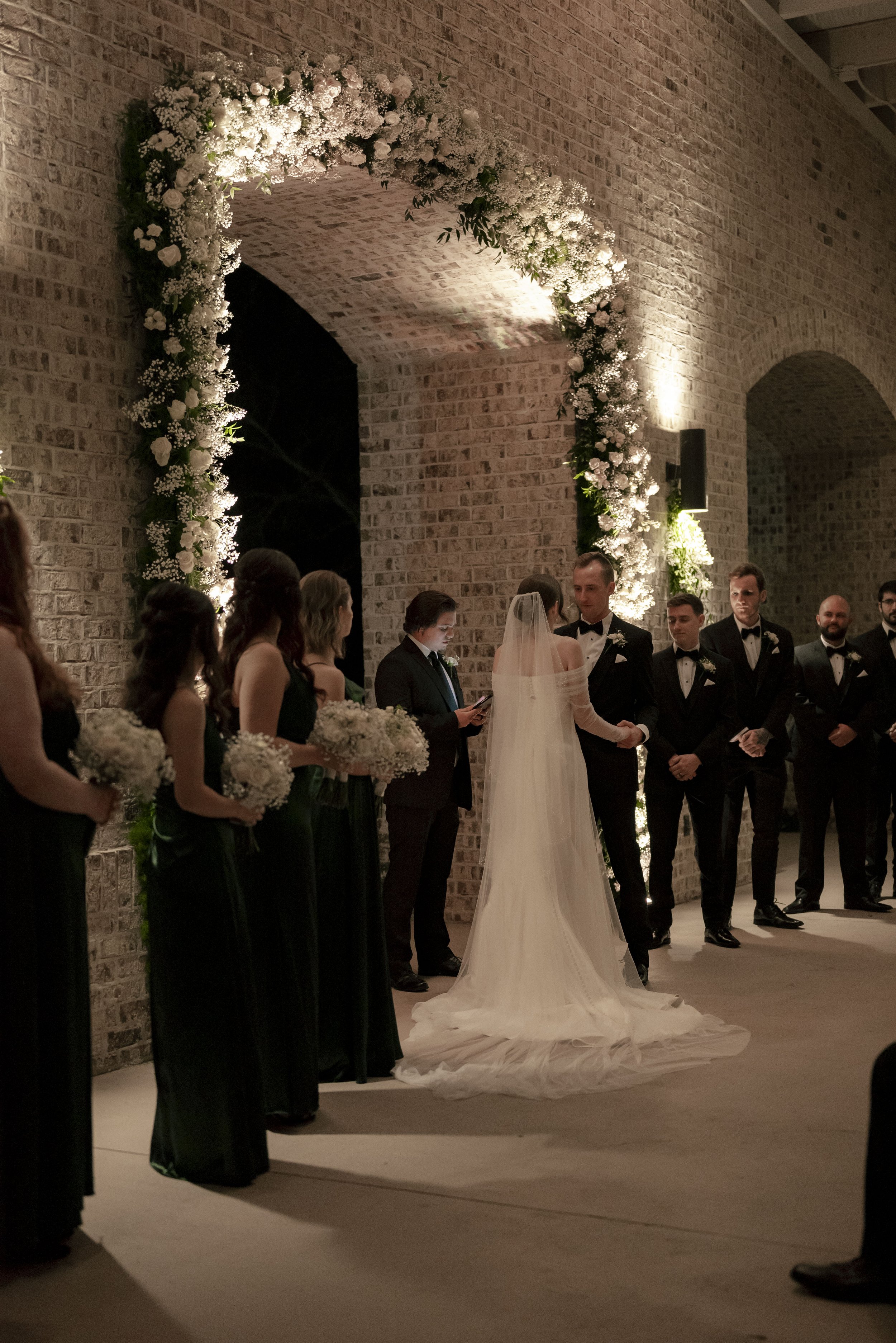 Bride and groom holding hands during wedding ceremony under floral arch with bridesmaids and groomsmen standing beside them in dimly lit venue with brick walls.