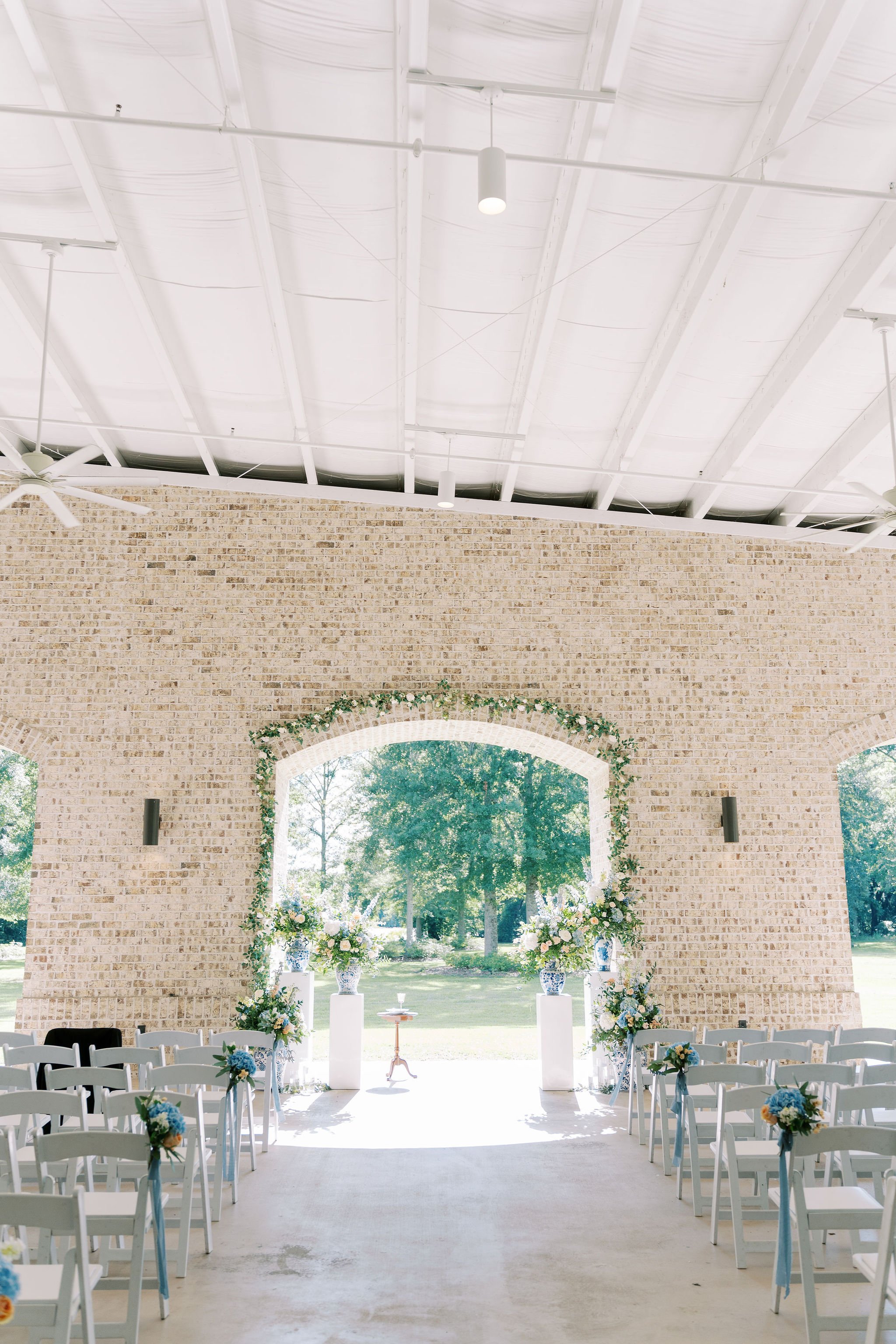 Indoor wedding ceremony setup with white chairs decorated with blue ribbons and floral arrangements, facing an archway adorned with greenery and flowers, with trees visible outside through large windows.