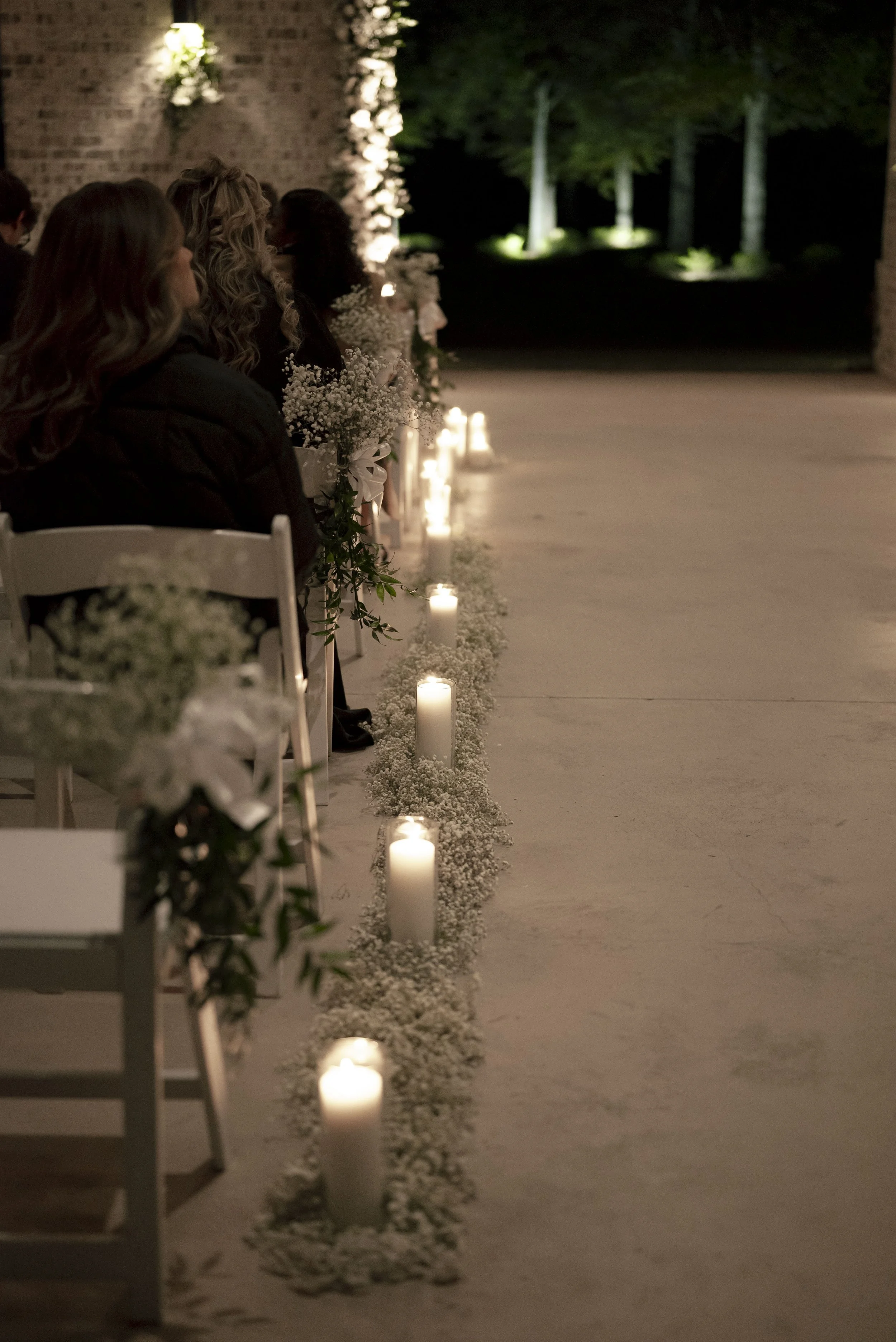 A nighttime outdoor wedding ceremony with a row of lit candles and flowers along the aisle, and guests sitting on white chairs facing forward.
