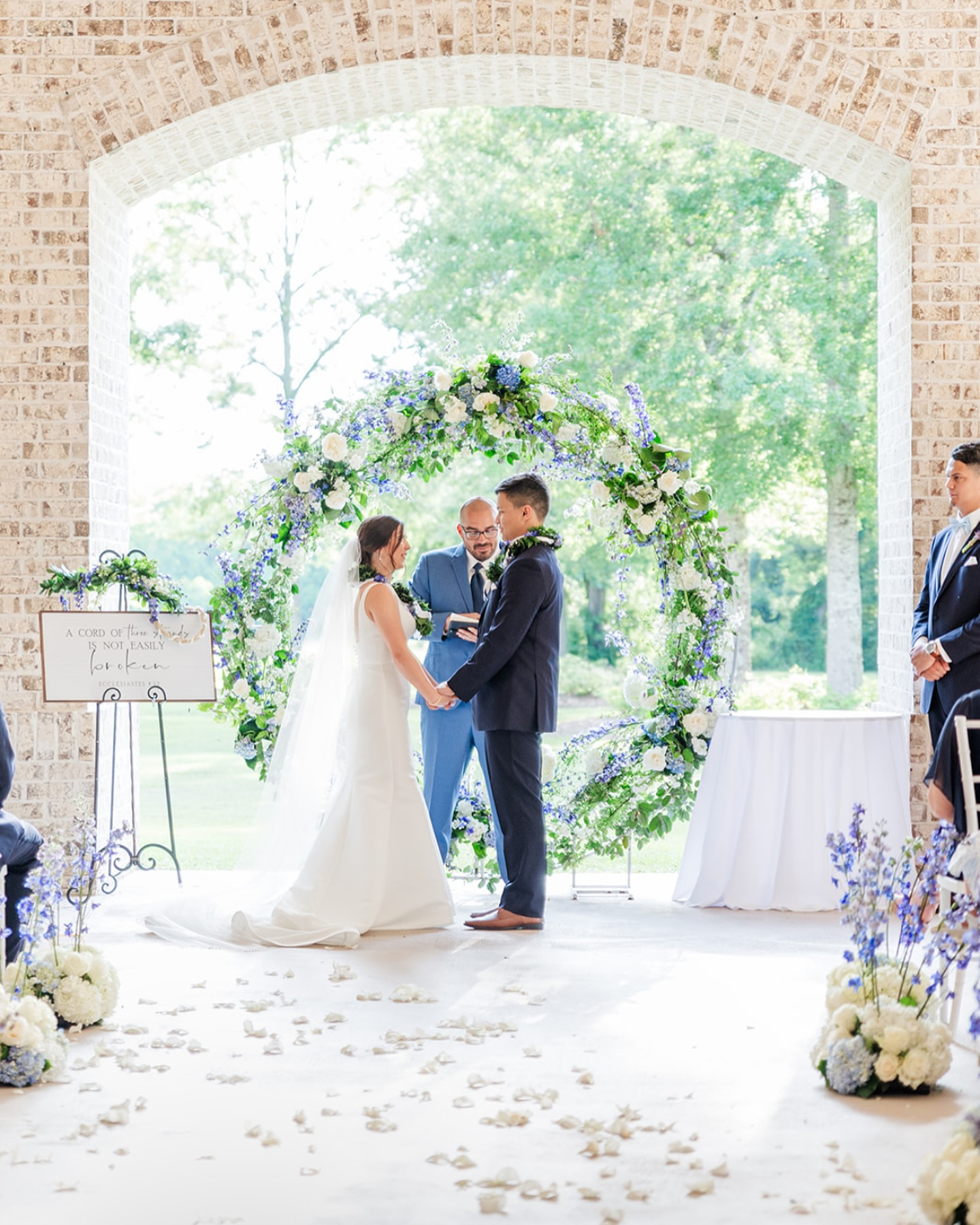 A wedding ceremony with a bride and groom holding hands under a floral arch, with an officiant officiating, in a venue with brick walls and large open windows showing trees outside.