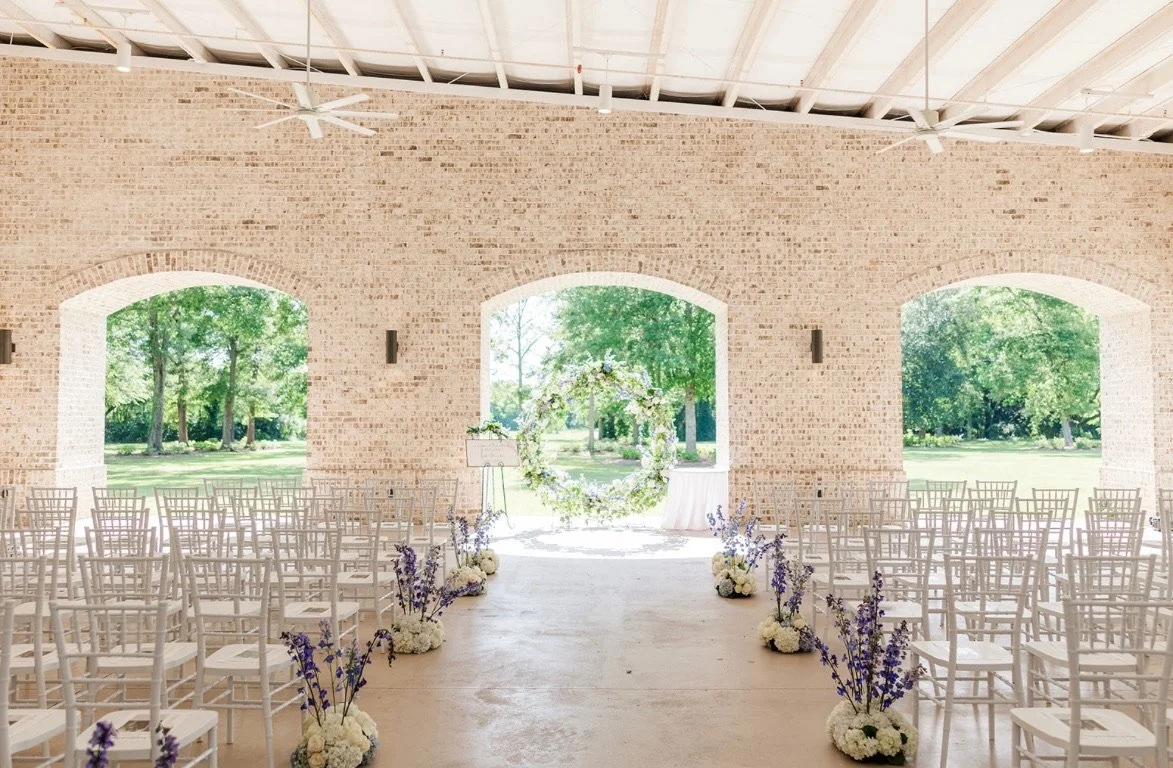 Interior of a decorated outdoor wedding venue with rows of white chairs, floral arrangements, and a floral arch at the altar, with open views of green trees outside.