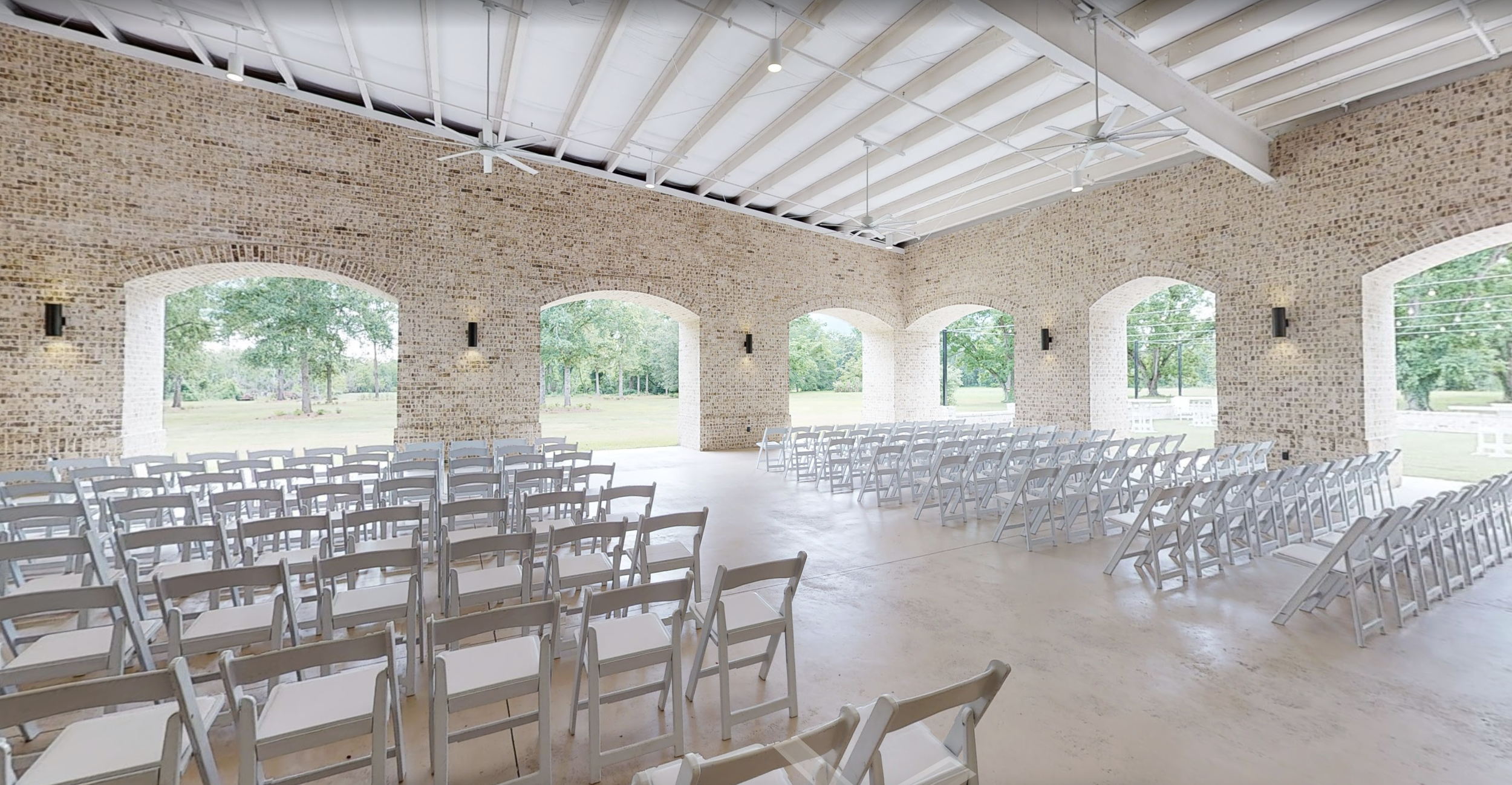 Empty indoor venue with white folding chairs arranged in rows, brick walls with large arched windows, outdoor view of trees and grass, ceiling fans, and industrial lighting fixtures.