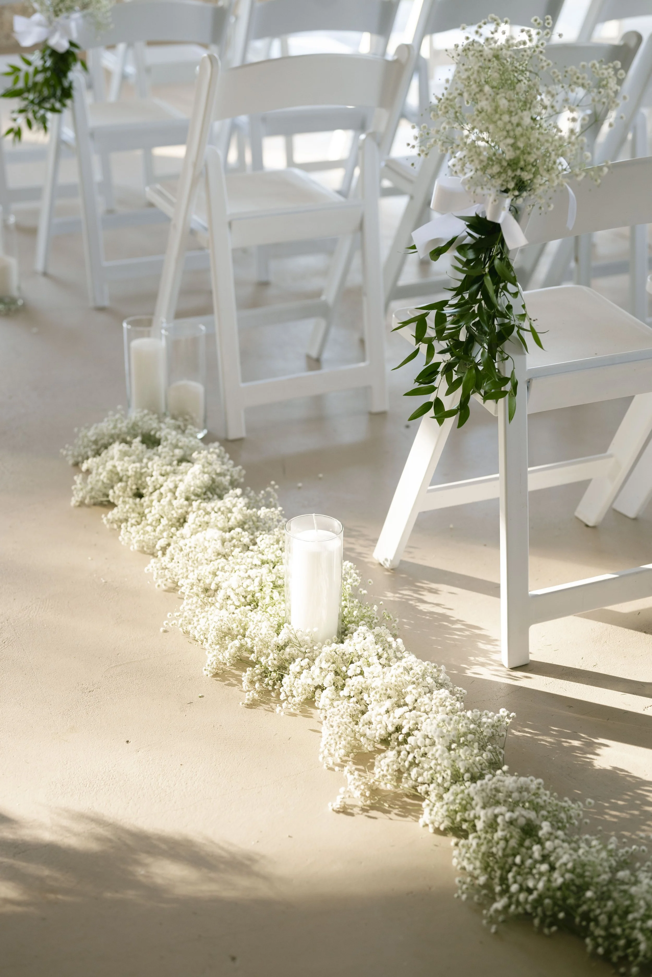 White chairs decorated with greenery and flowers, white candles in glass holders, and a floral arrangement for a wedding ceremony or event.