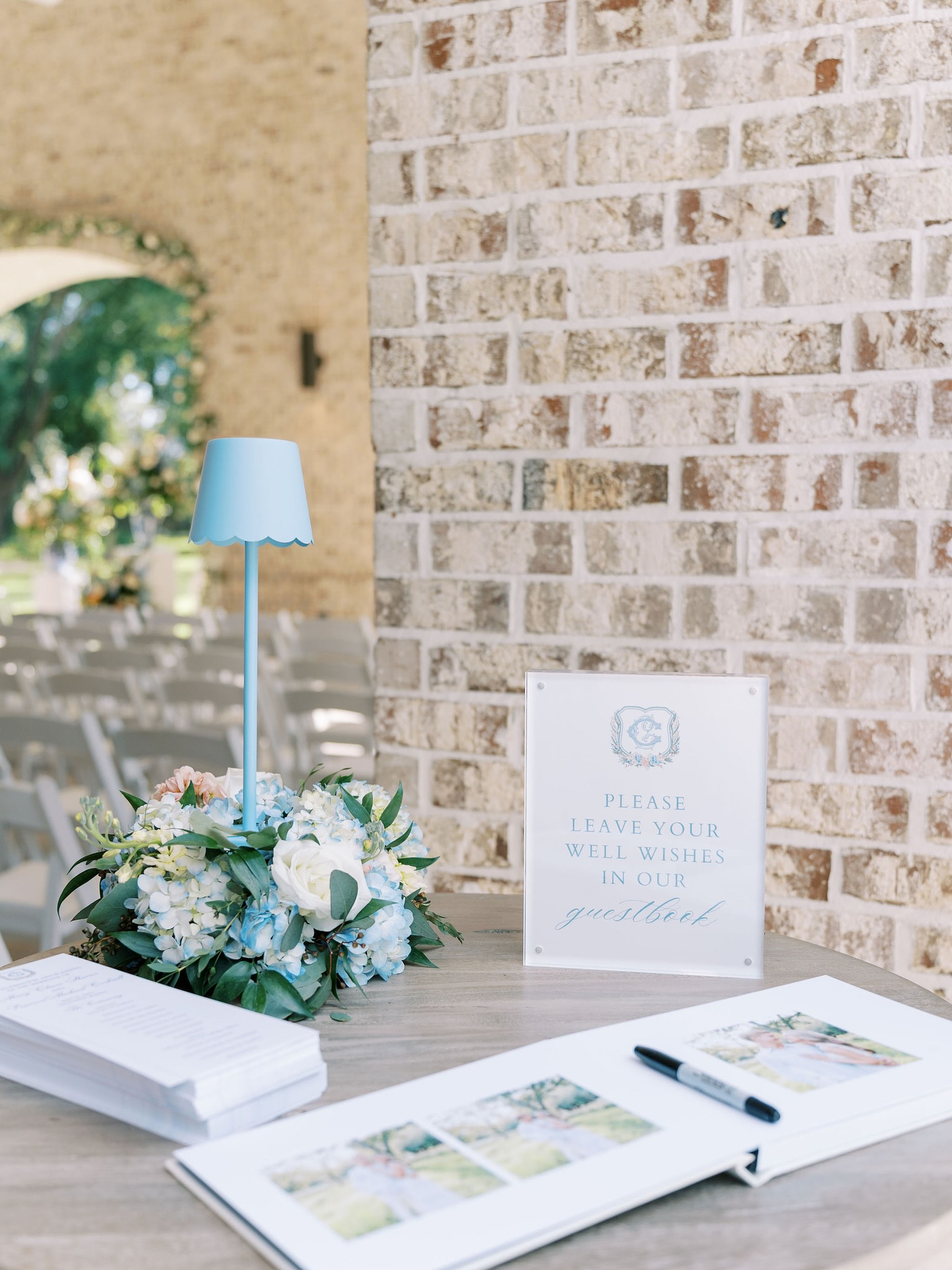 A guestbook table at a wedding with a sign instructing guests to leave well wishes, a floral centerpiece, a pen, and photo pages for guests to sign.