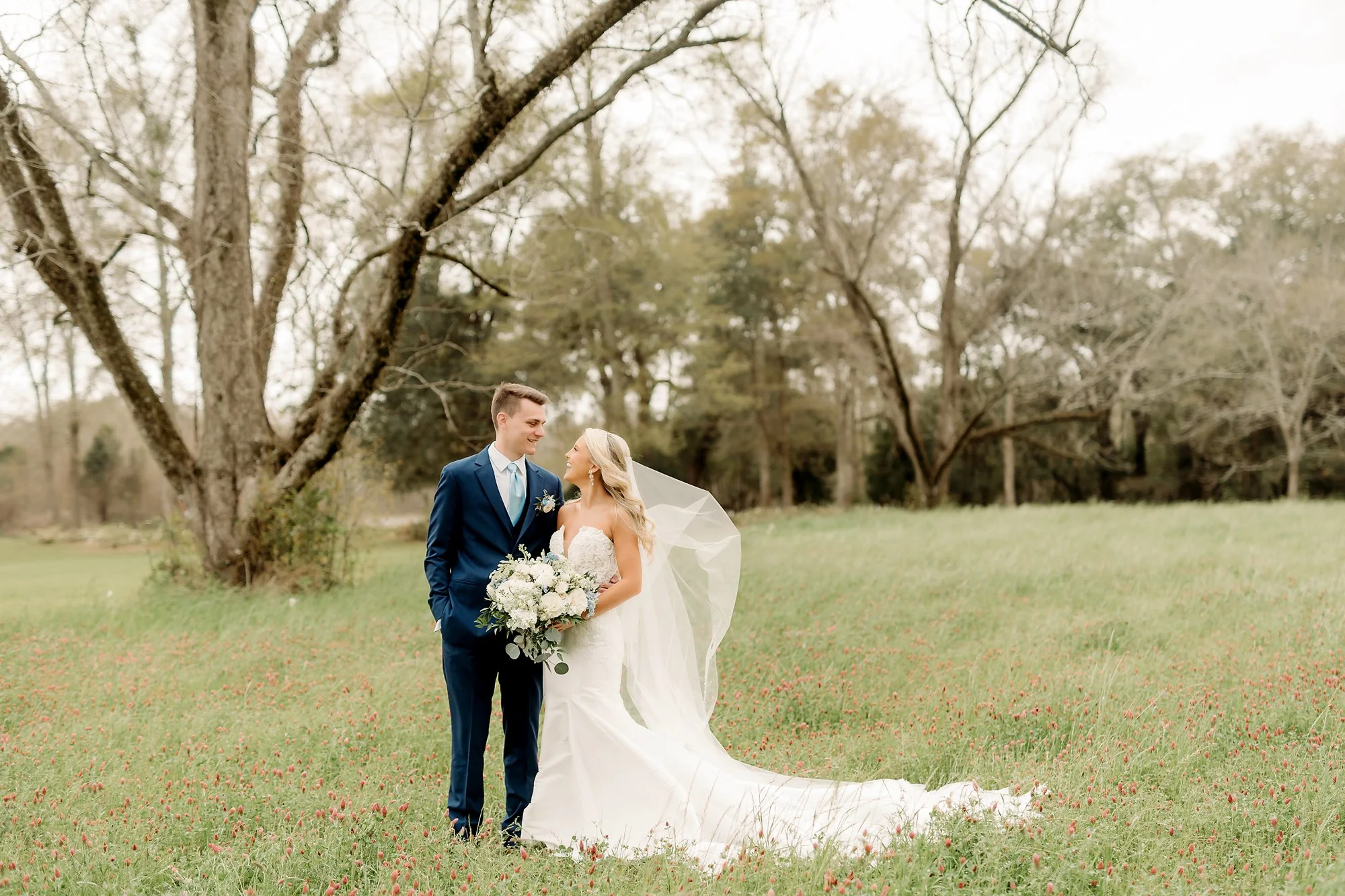 Bride and groom standing together on a grassy field with trees in the background, the bride holding a bouquet of flowers, both smiling at each other.