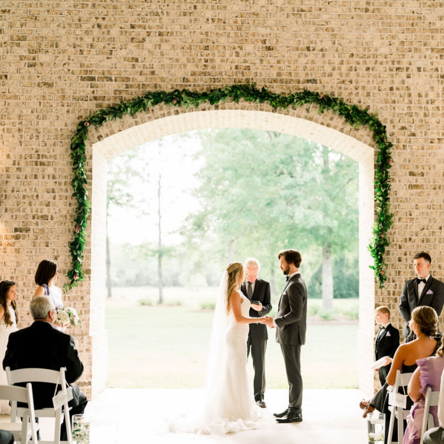 Bride and groom exchanging vows during outdoor wedding ceremony in front of officiant, with guests seated on chairs on both sides, brick arch decorated with greenery behind them.