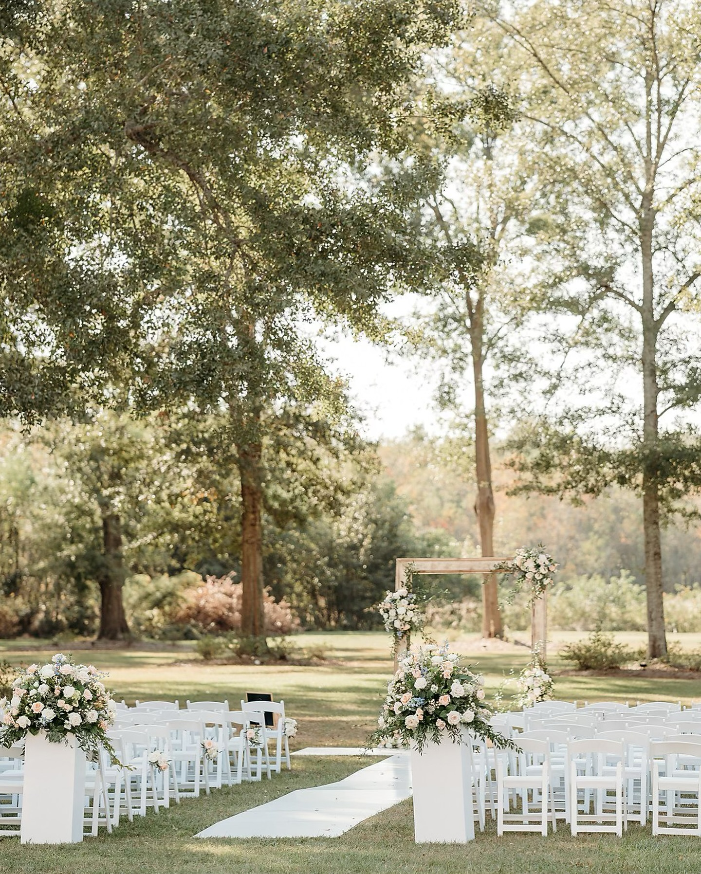 Outdoor wedding setup with white chairs, floral arrangements, and a wooden arch on a grassy area surrounded by trees.