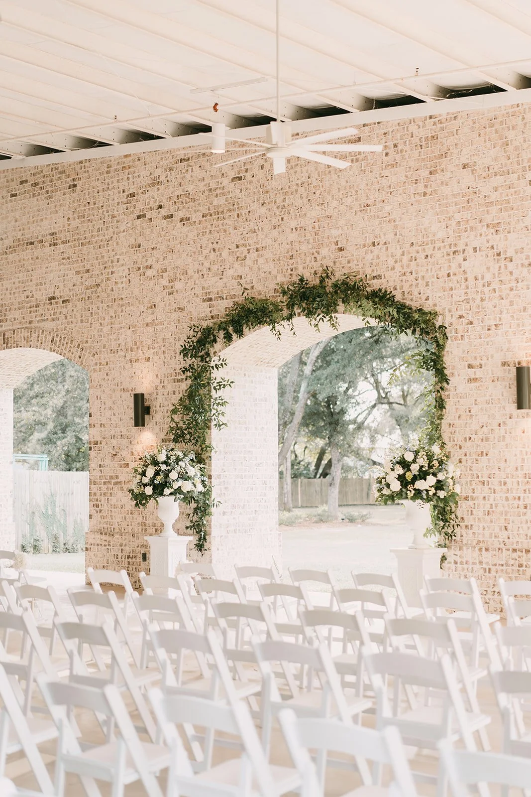 Wedding ceremony setup with white chairs, floral arrangements, and a greenery arch on a brick wall inside a bright space with large arched windows.
