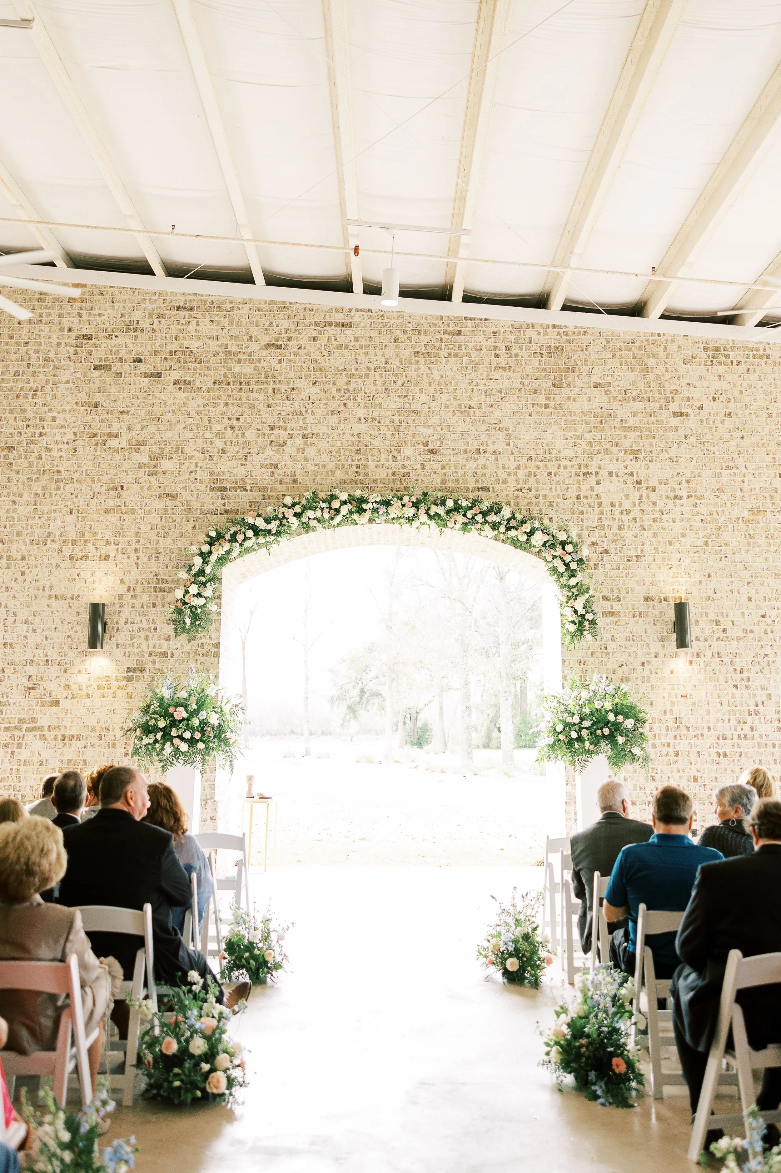 Indoor wedding ceremony with guests seated on either side of an aisle decorated with floral arrangements. An arch with flowers marks the altar, leading to an outdoor view of trees and bright natural light.