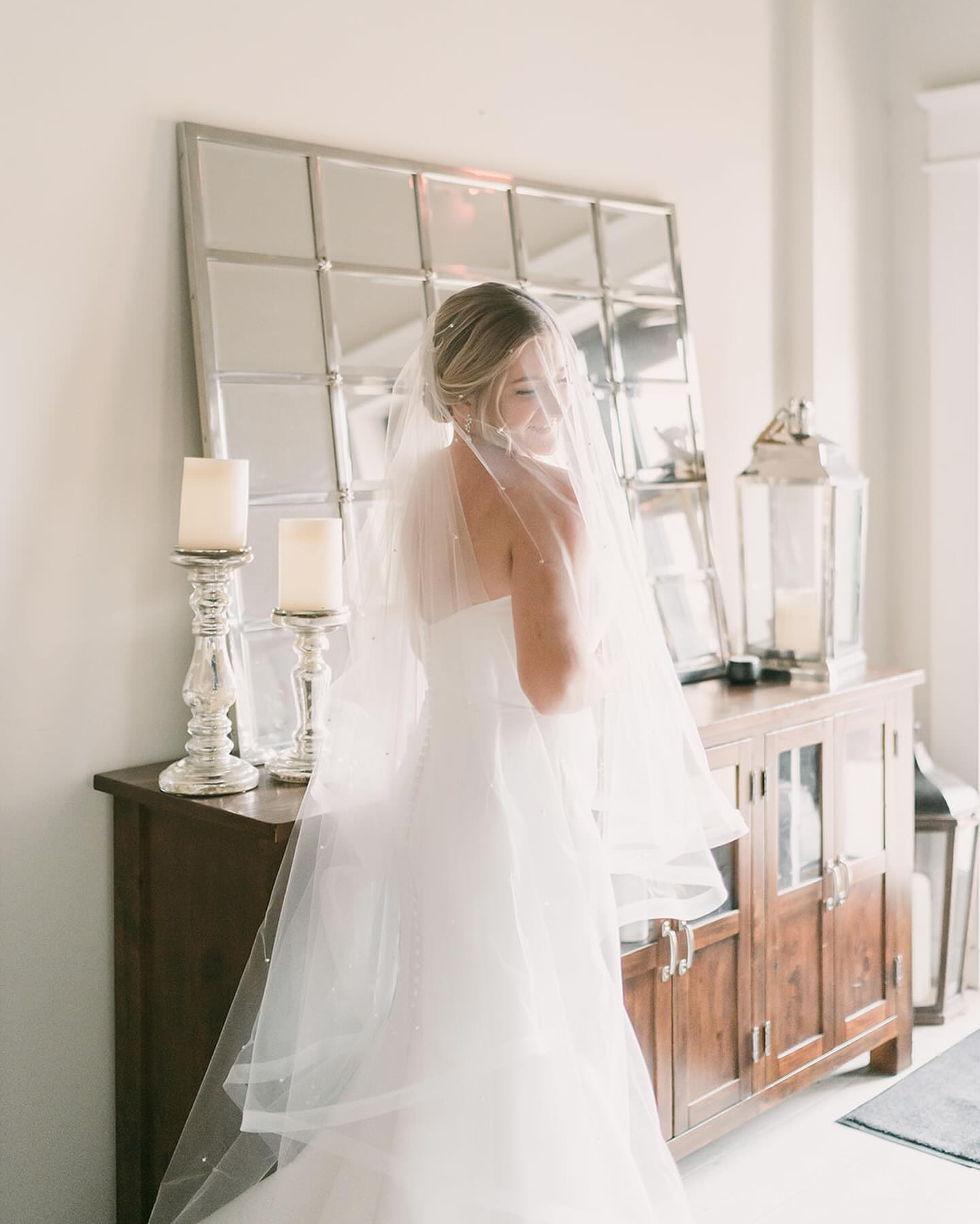 Bride standing and smiling in a wedding dress, veil, and earrings in a bright room with a mirror, candles, and wooden furniture.