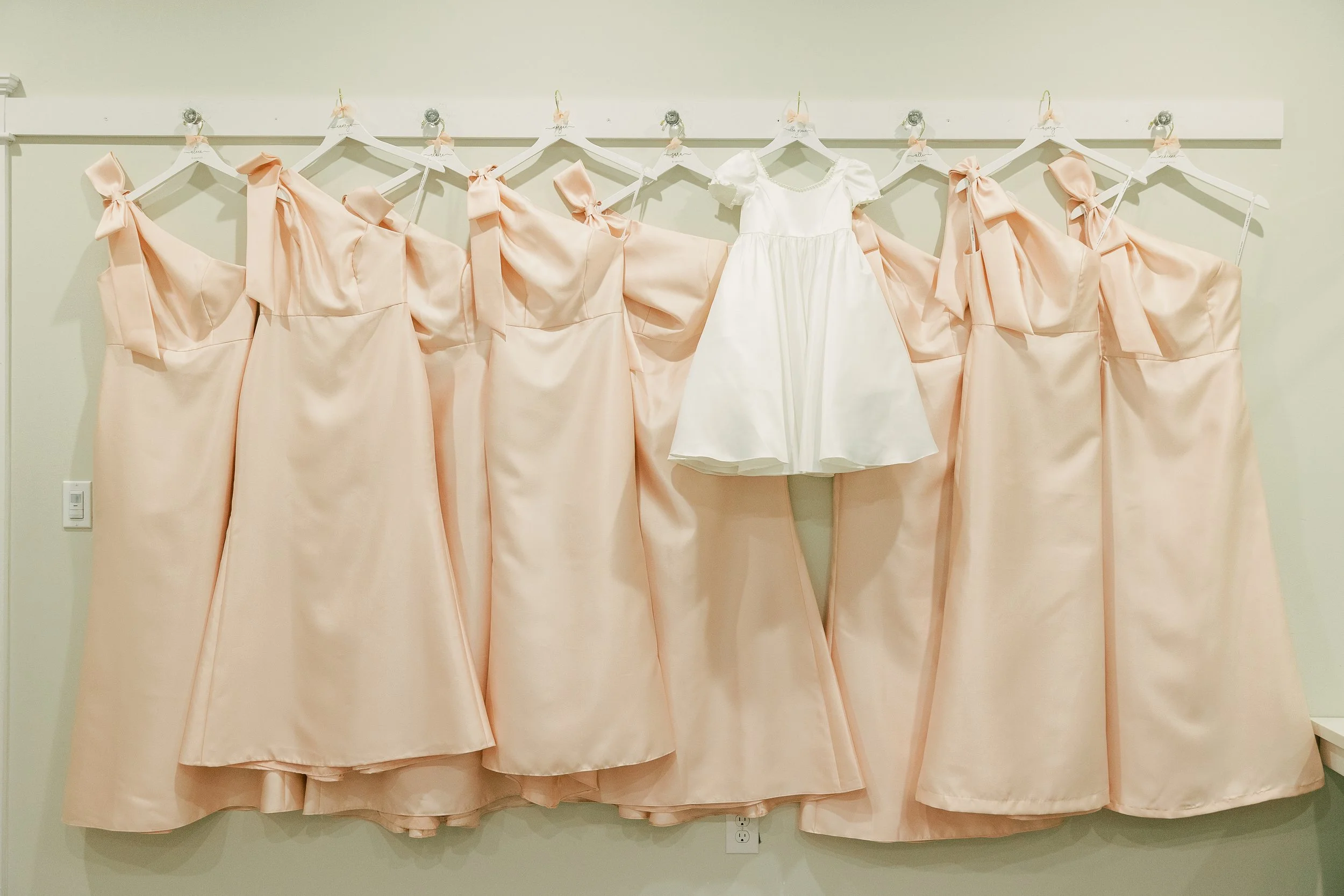 Seven peach-colored dresses with bow details hanging on a wall, with one white dress in the center.
