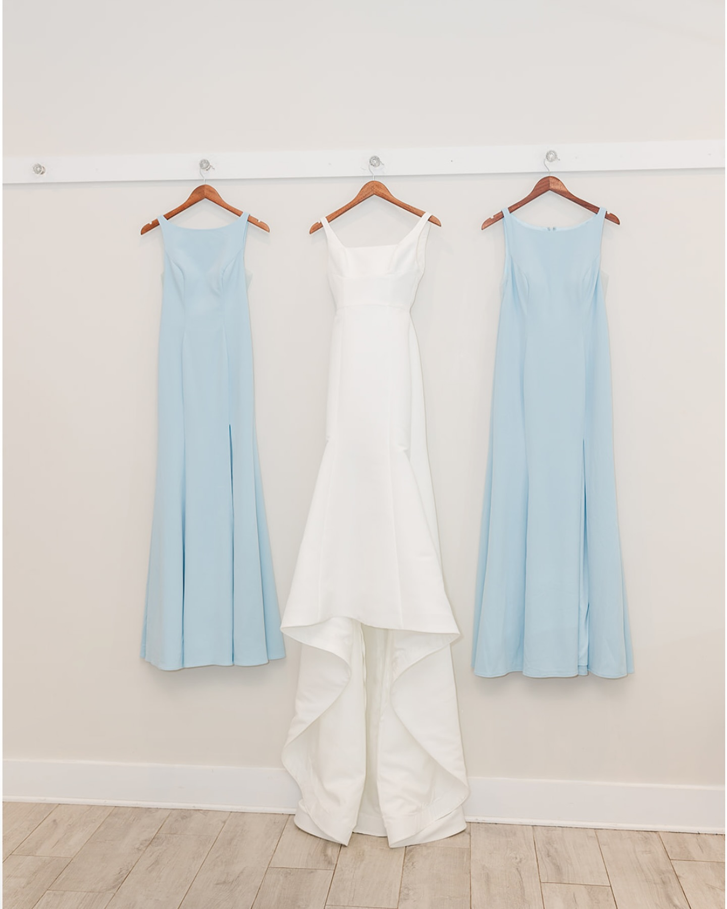 A white wedding dress hangs in the center on a wooden hanger, flanked by two blue bridesmaid dresses on either side, all hanging on a white wall with a thin horizontal rail.