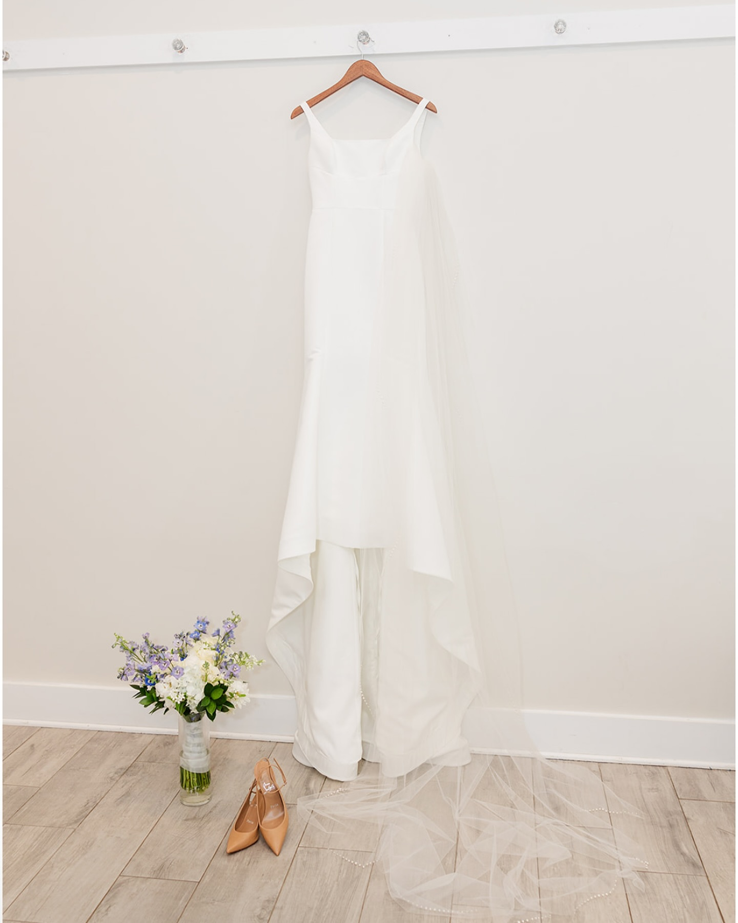 White wedding dress on a hanger, flower bouquet, beige shoes, and veil on light wooden floor