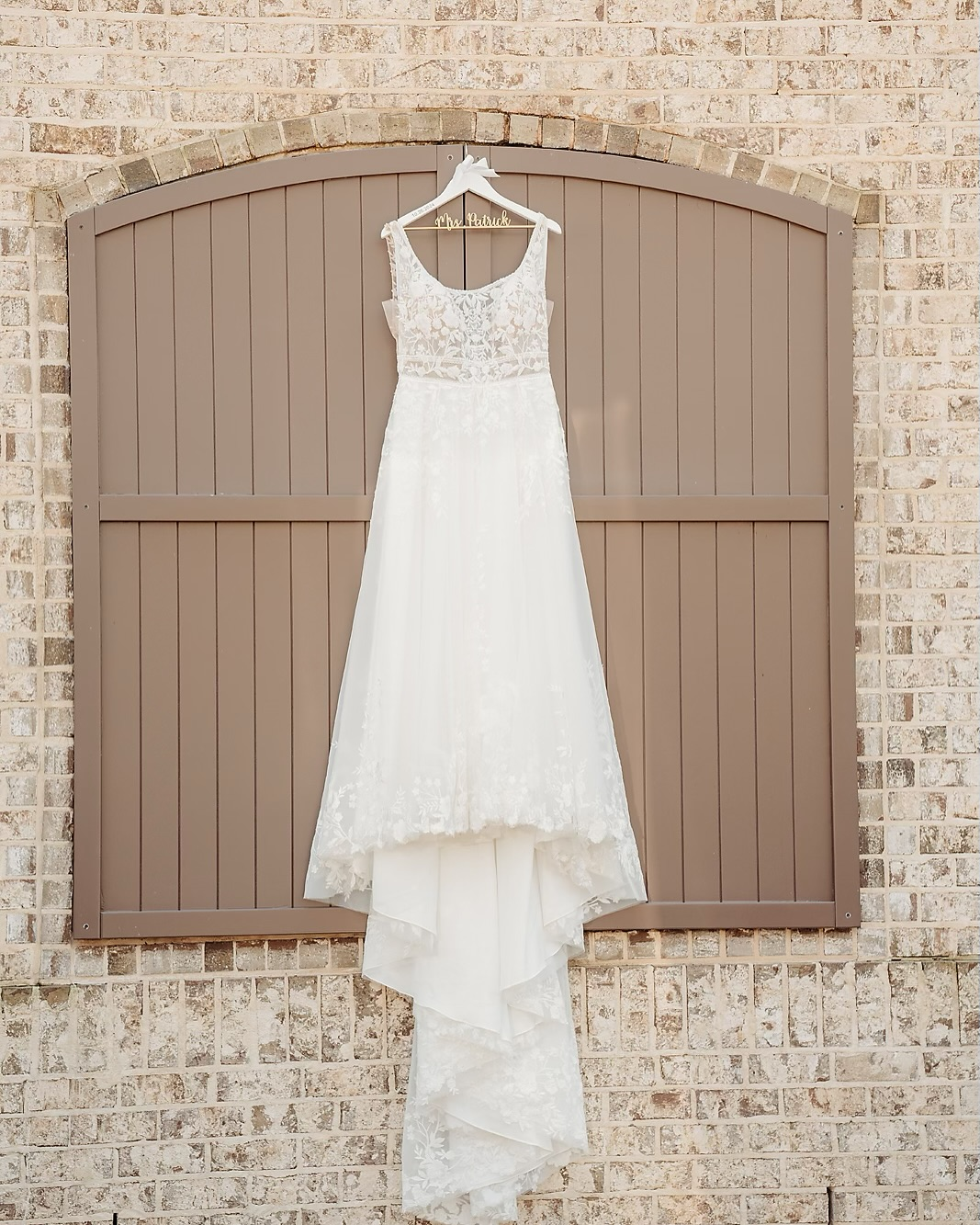 White wedding dress hanging on a hanger with a wooden name tag that says 'Mrs. Patrick' on a brown fence with a brick wall background.