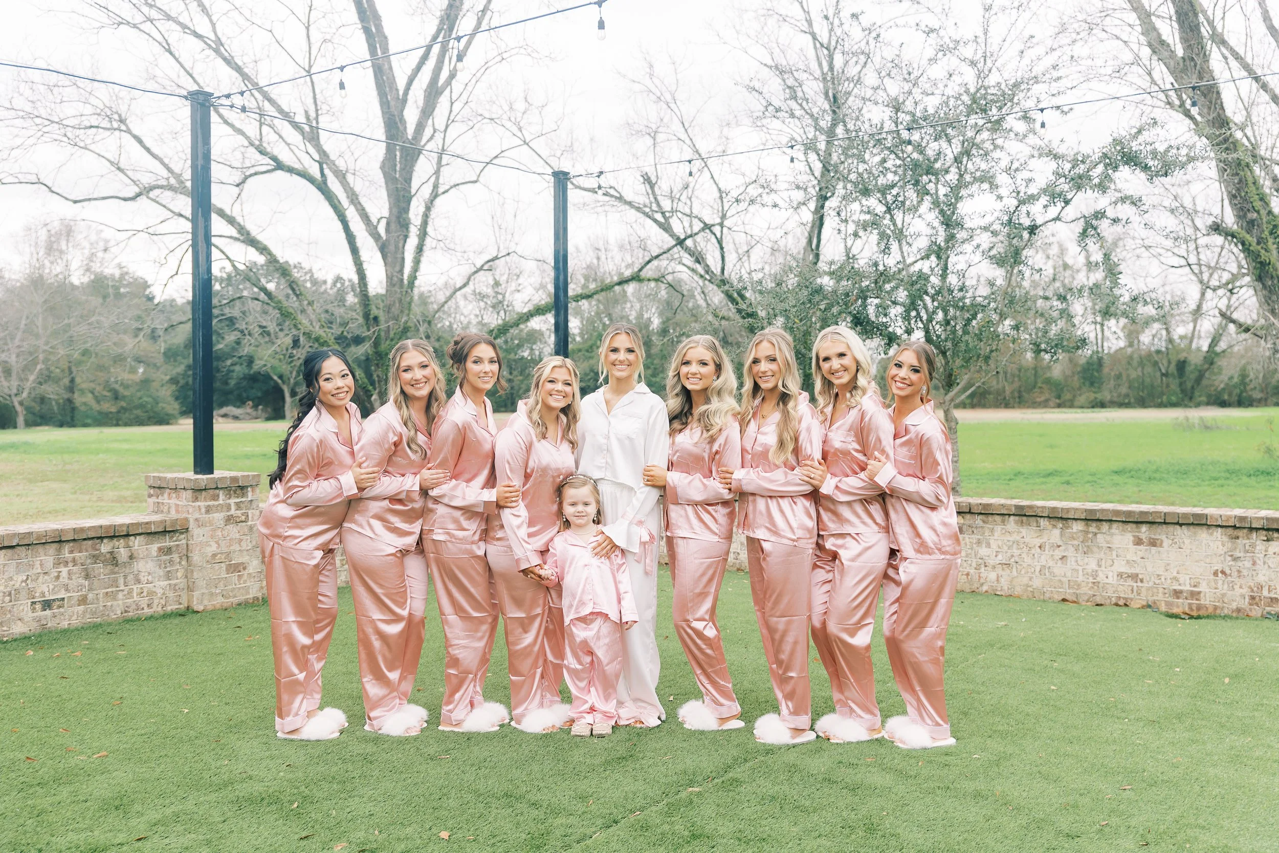 A group of ten women and one young girl standing outdoors on grass, wearing matching pink satin pajamas and fluffy slippers, smiling for a photo during daytime.