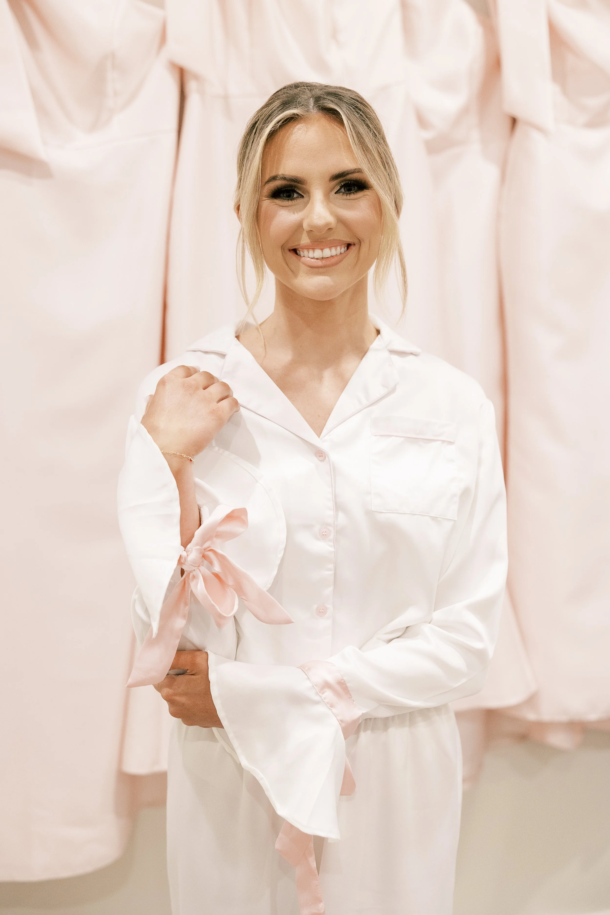 Young woman smiling, wearing white satin pajamas with pink ribbon details, standing in front of hanging pajamas.