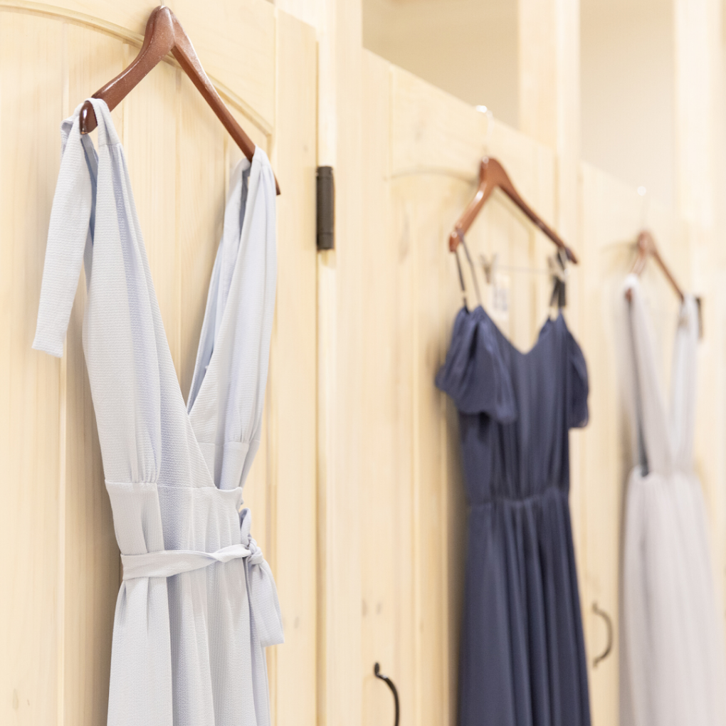 Hanging dresses, white, navy, and light-colored, on wooden hangers against light wood-paneled wall.