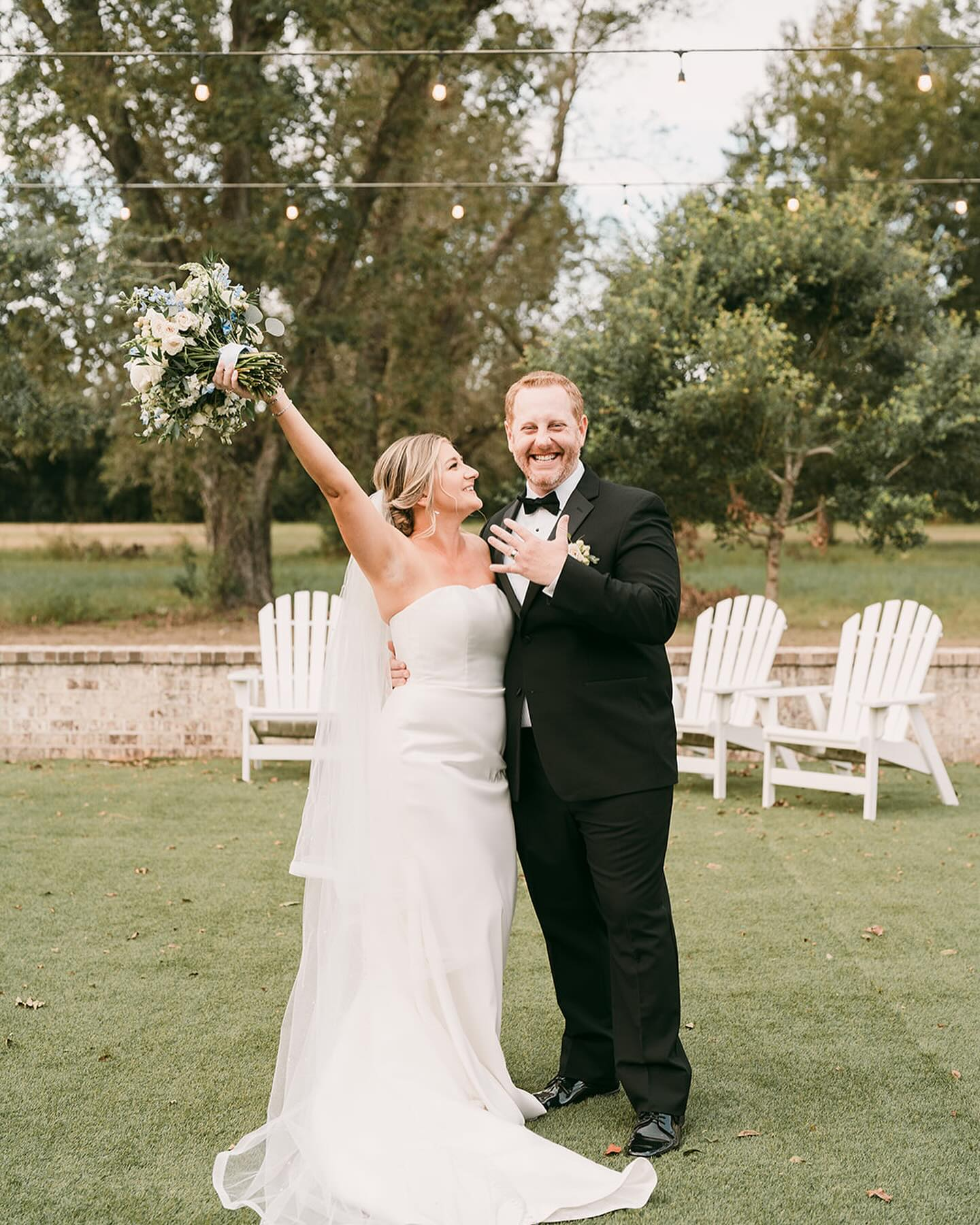 Bride and groom celebrating outdoors on their wedding day, with the bride holding a bouquet of flowers and the groom smiling in a black tuxedo, in a garden setting with trees and white Adirondack chairs.
