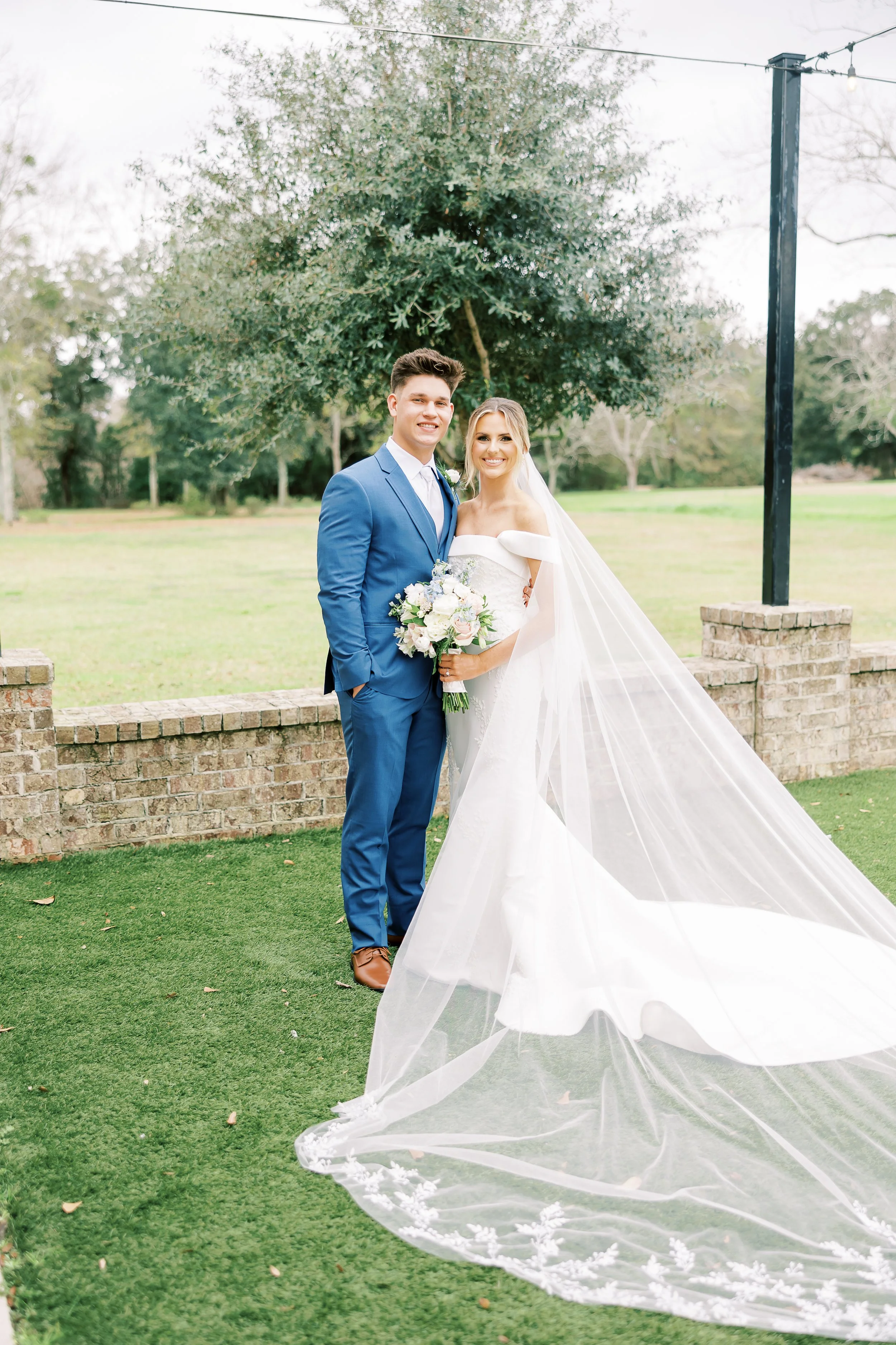 A bride and groom standing outdoors on a grassy area, with a tree and a brick wall behind them, celebrating their wedding day. The bride is in a white wedding dress with a long veil, holding a bouquet, and the groom is in a blue suit with a white shi