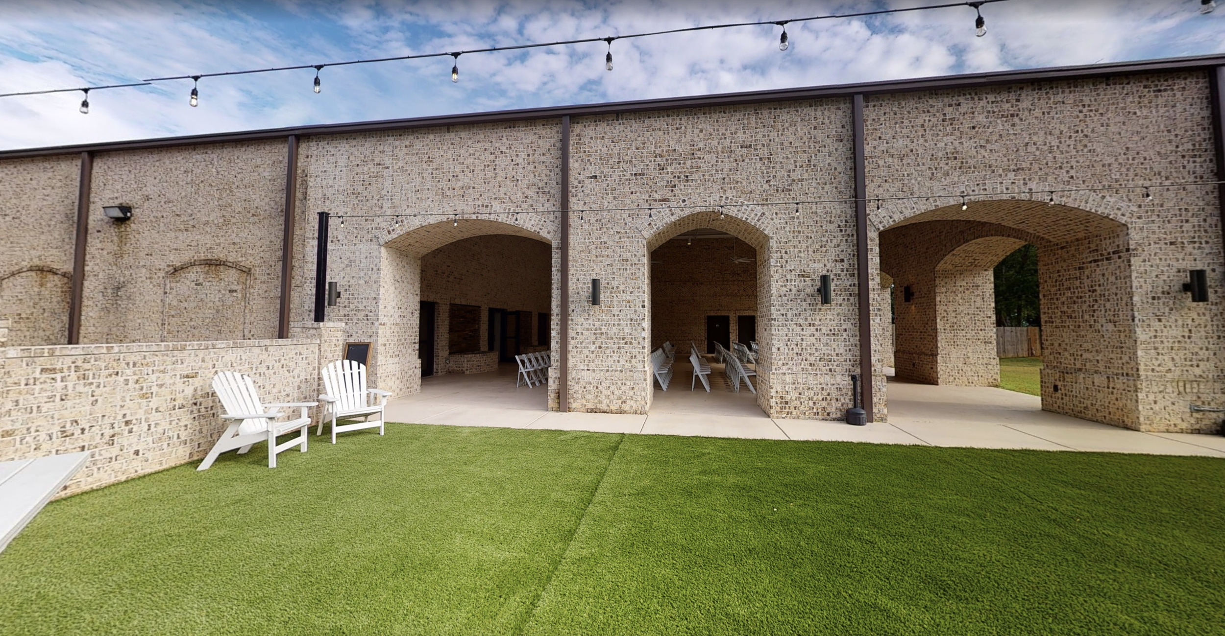 Outdoor patio area with brick arches, white chairs, and string lights above, with a grass lawn in the foreground and a partly cloudy sky.