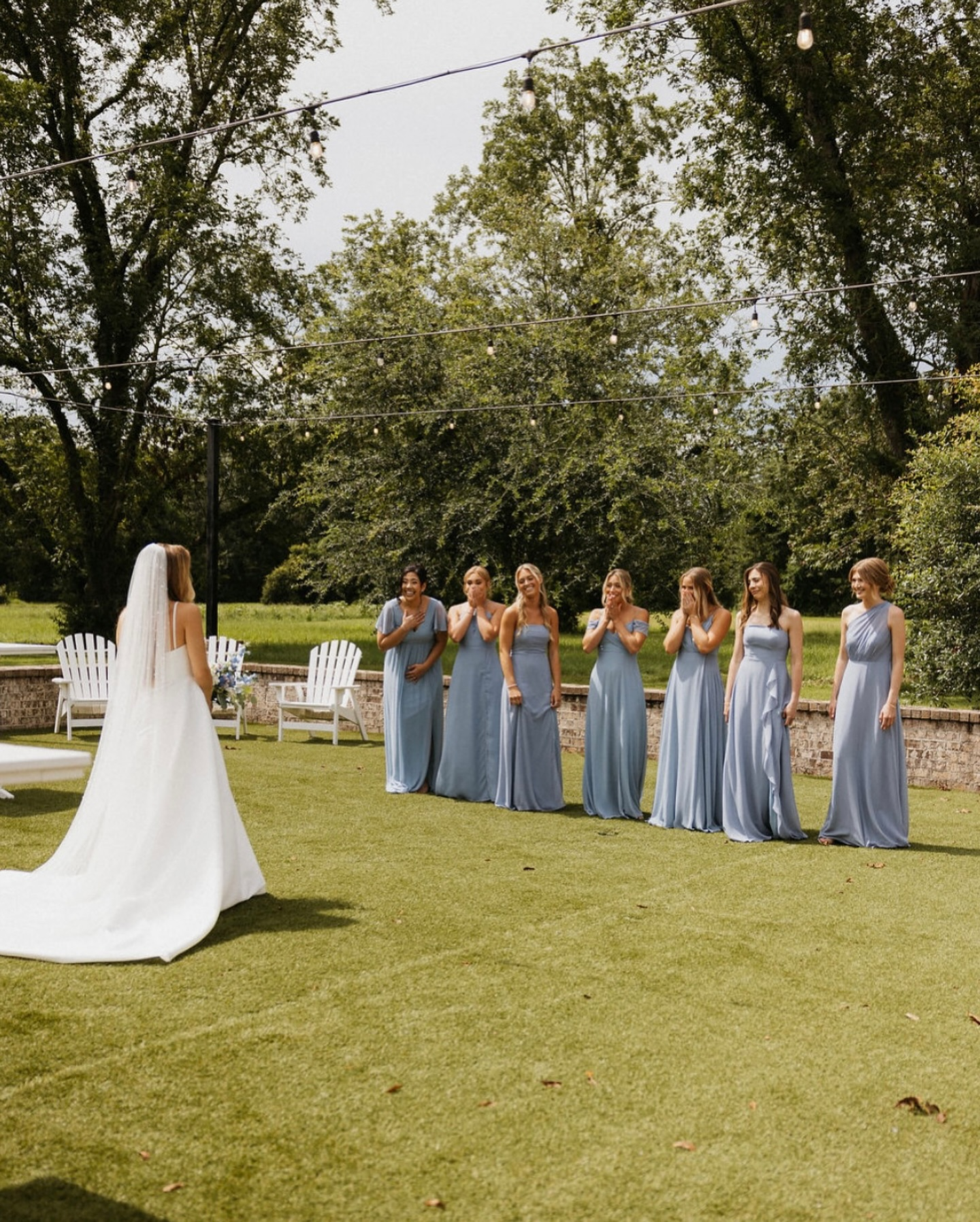 Bride and six bridesmaids standing outdoors on a grassy area, with trees and string lights overhead, during a wedding ceremony.