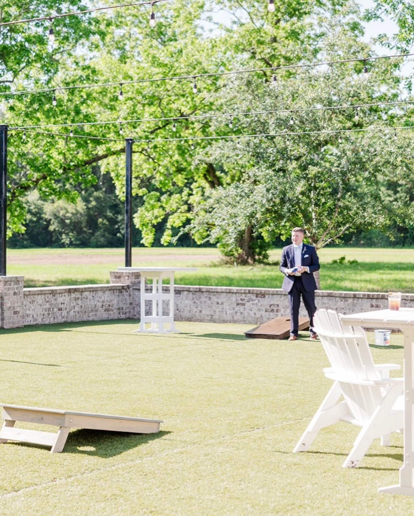 Man in a suit speaking outdoors at a wedding or event, with green trees, string lights, and white chairs in the background.