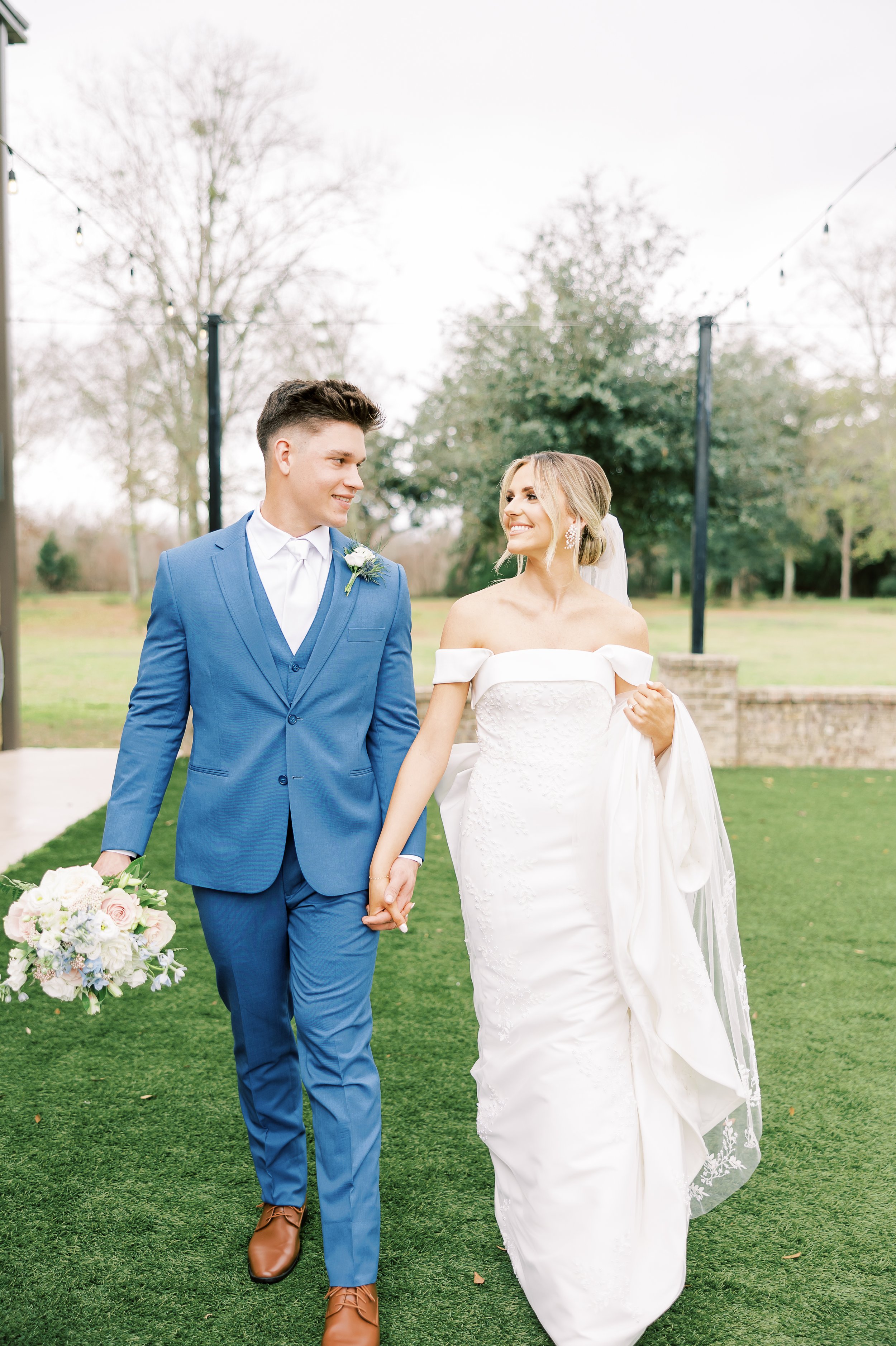 A bride and groom walking hand in hand outdoors, smiling at each other, with trees and string lights in the background.