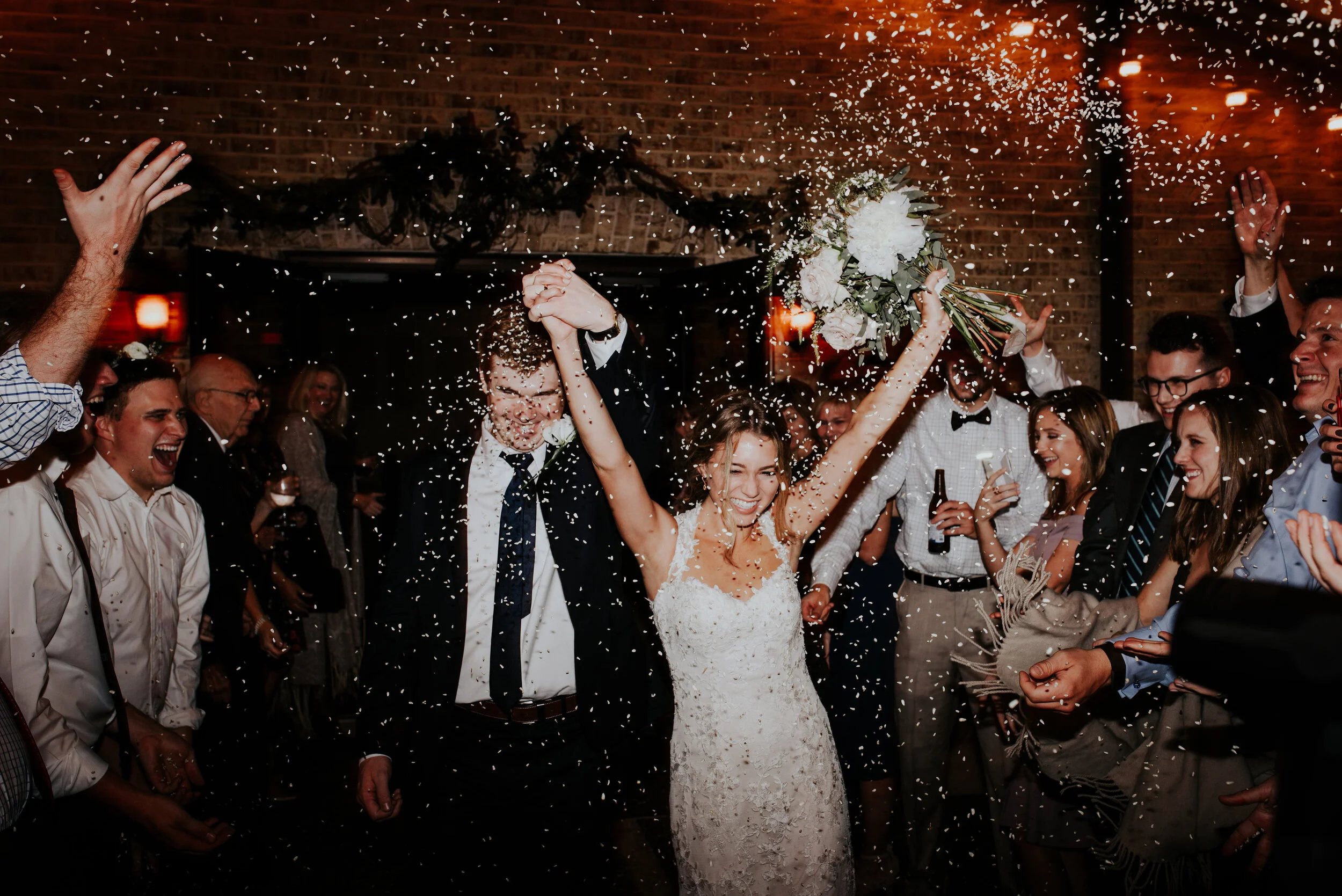 Bride and groom celebrating wedding with guests while throwing confetti, bride holding bouquet, indoor reception with warm lighting and brick walls.