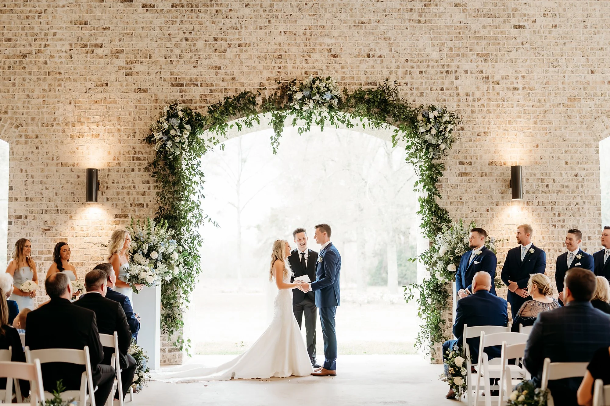 A wedding ceremony with a bride and groom exchanging vows under a floral arch, surrounded by bridesmaids and groomsmen, with seated guests watching indoors against a brick wall.