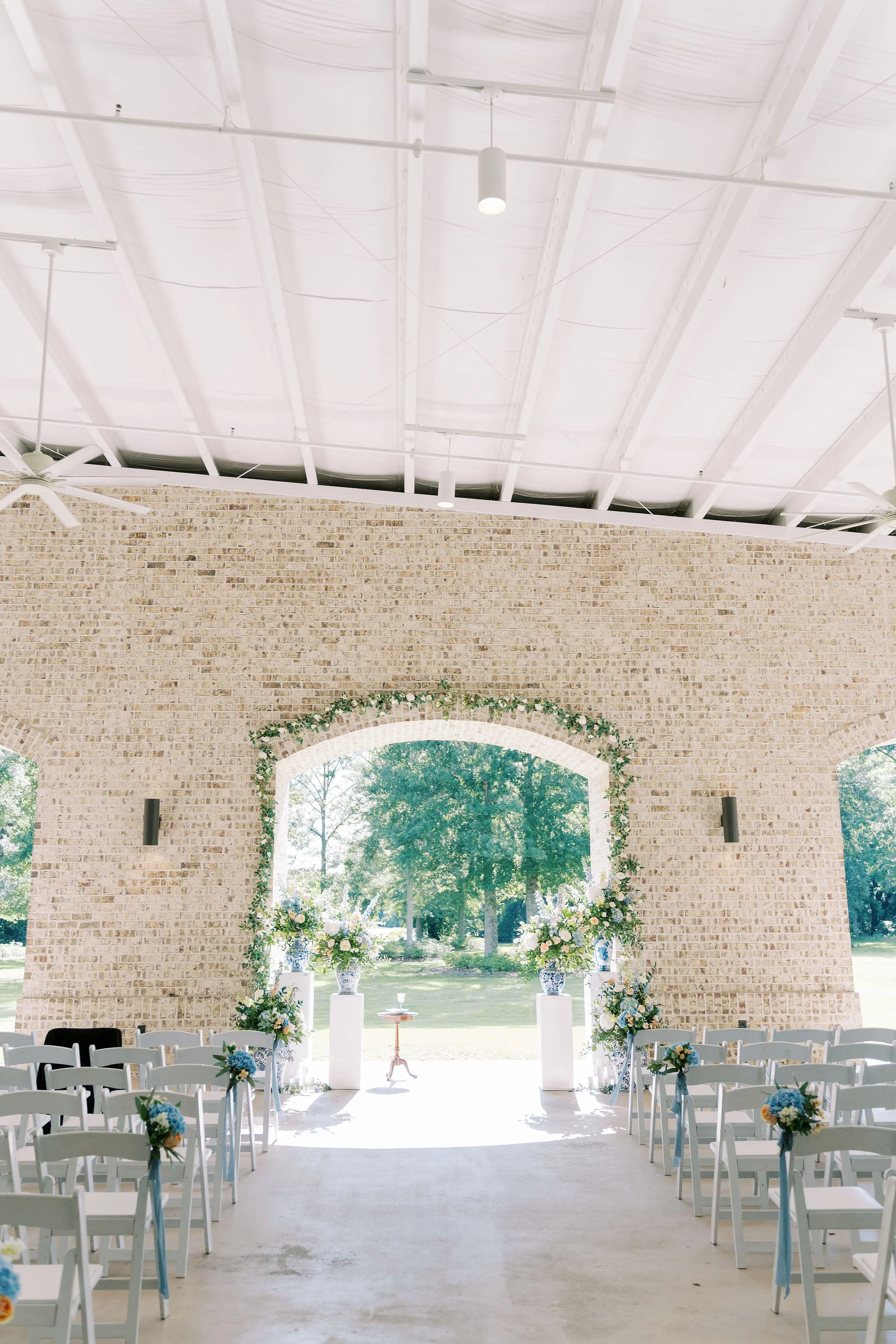 Wedding ceremony setup inside a modern, airy venue with a brick wall backdrop decorated with floral arrangements, rows of white chairs, and an open view of a green outdoor landscape.