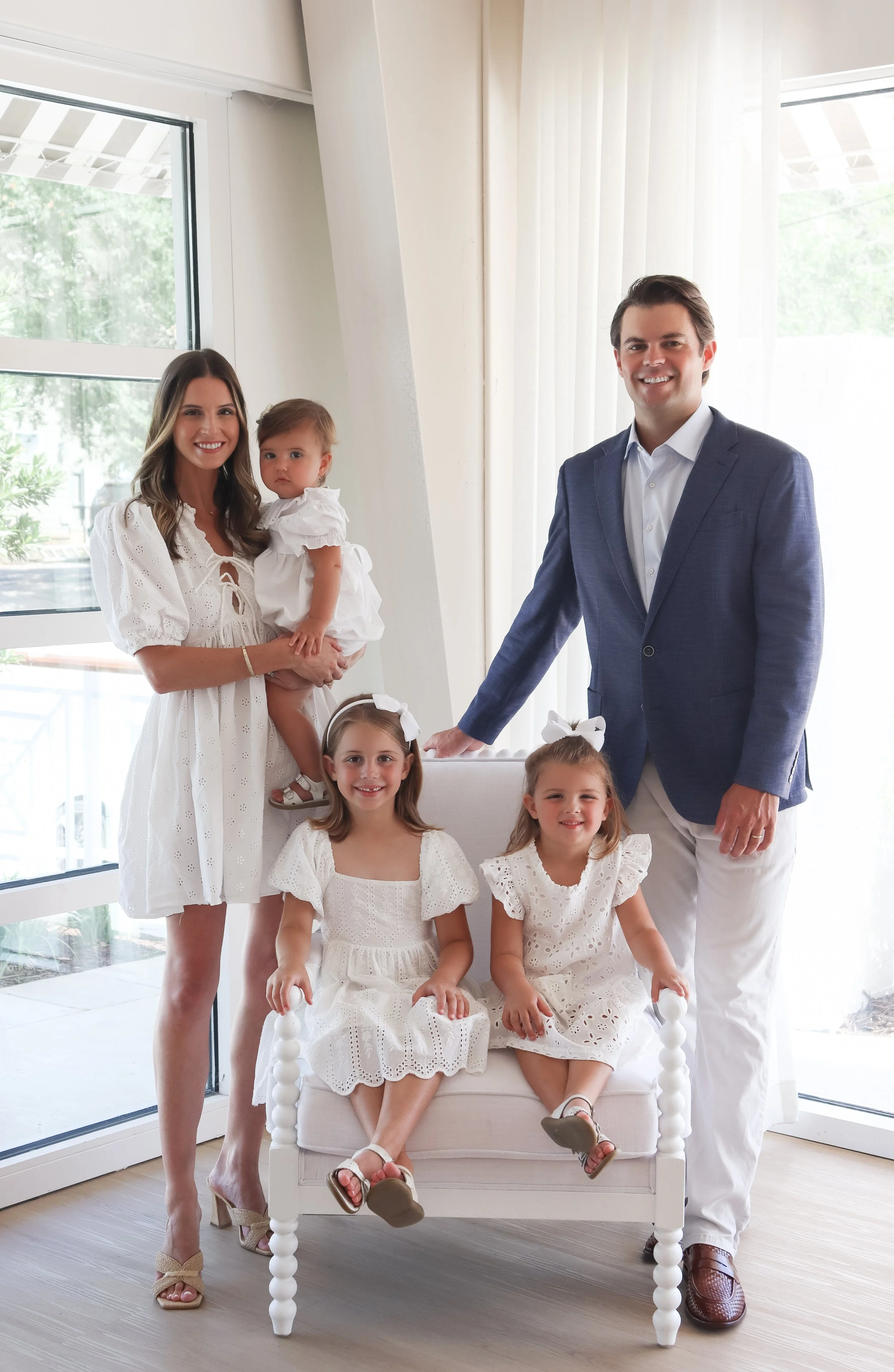 Family portrait of a woman, a man, and three young girls in white dresses, in a well-lit room with large windows.