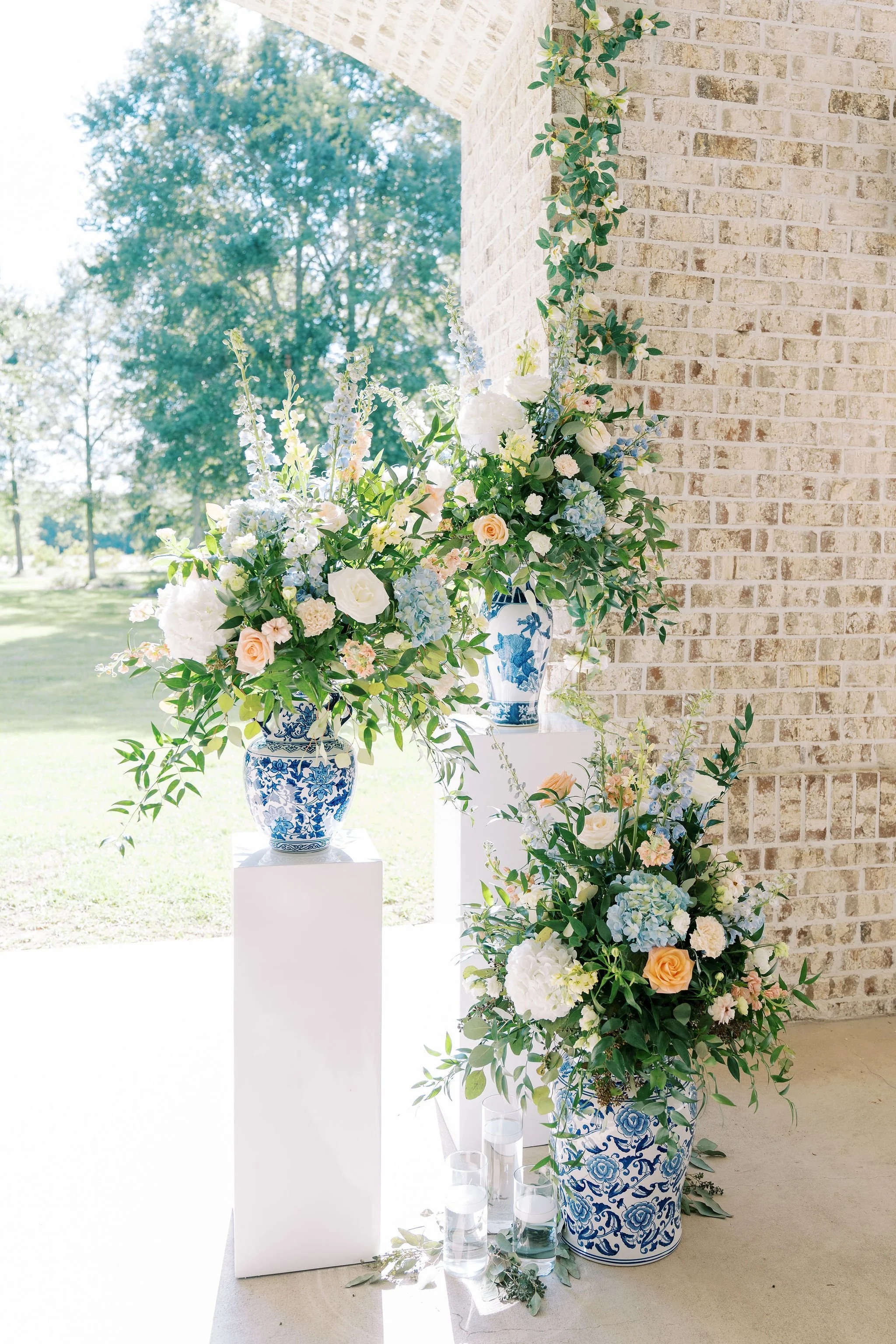 Arrangement of white, pastel pink, and light blue flowers in blue and white patterned vases, with candles at the base, near a brick wall with greenery outside.