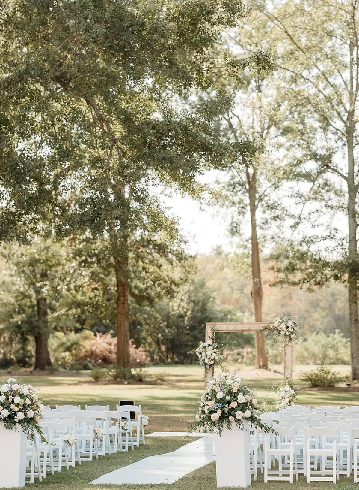 Outdoor wedding setup with white chairs, floral arrangements, and a wooden arch under trees.