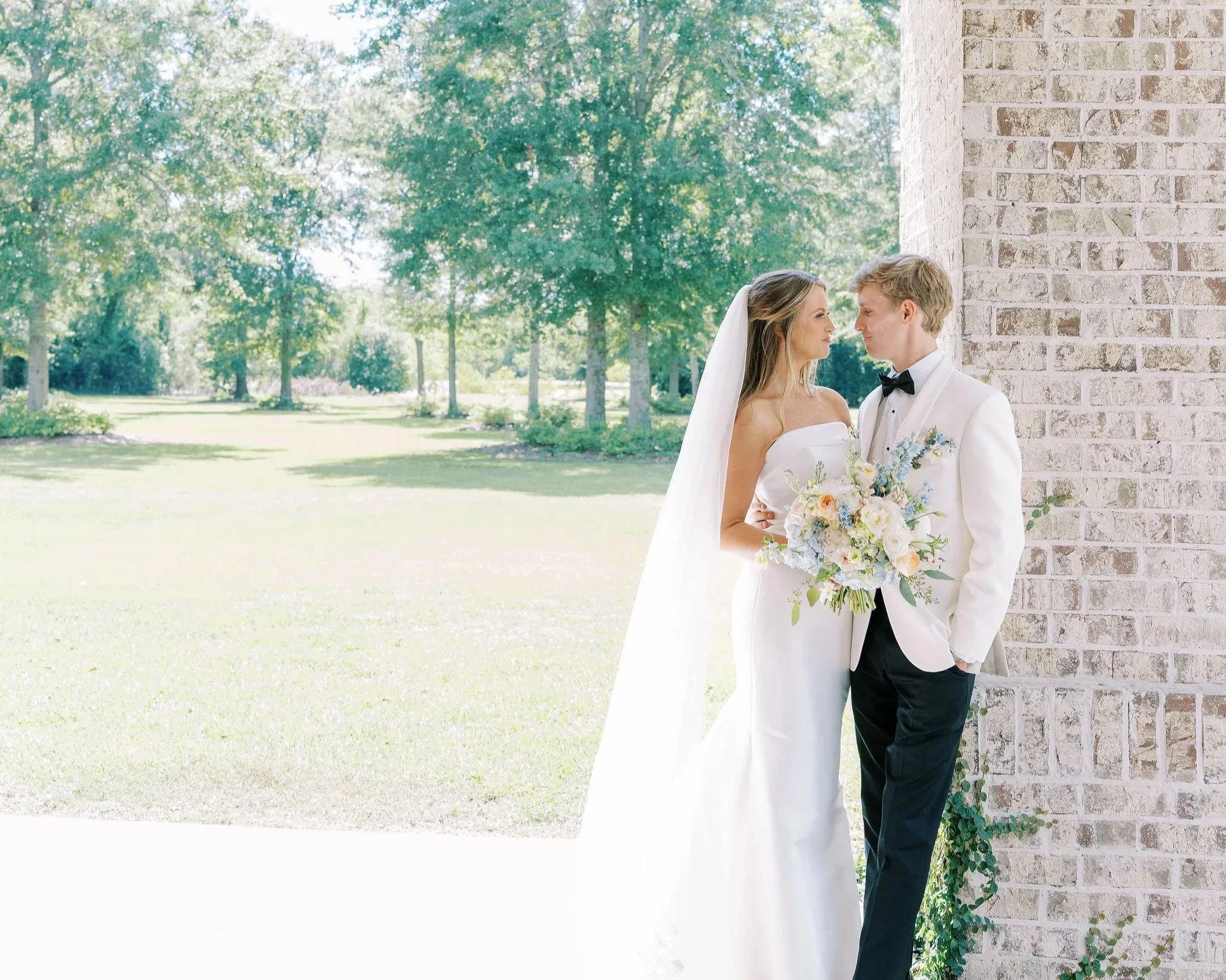A bride and groom in wedding attire standing near a brick wall outdoors, holding a bouquet of flowers, with trees and grass in the background.