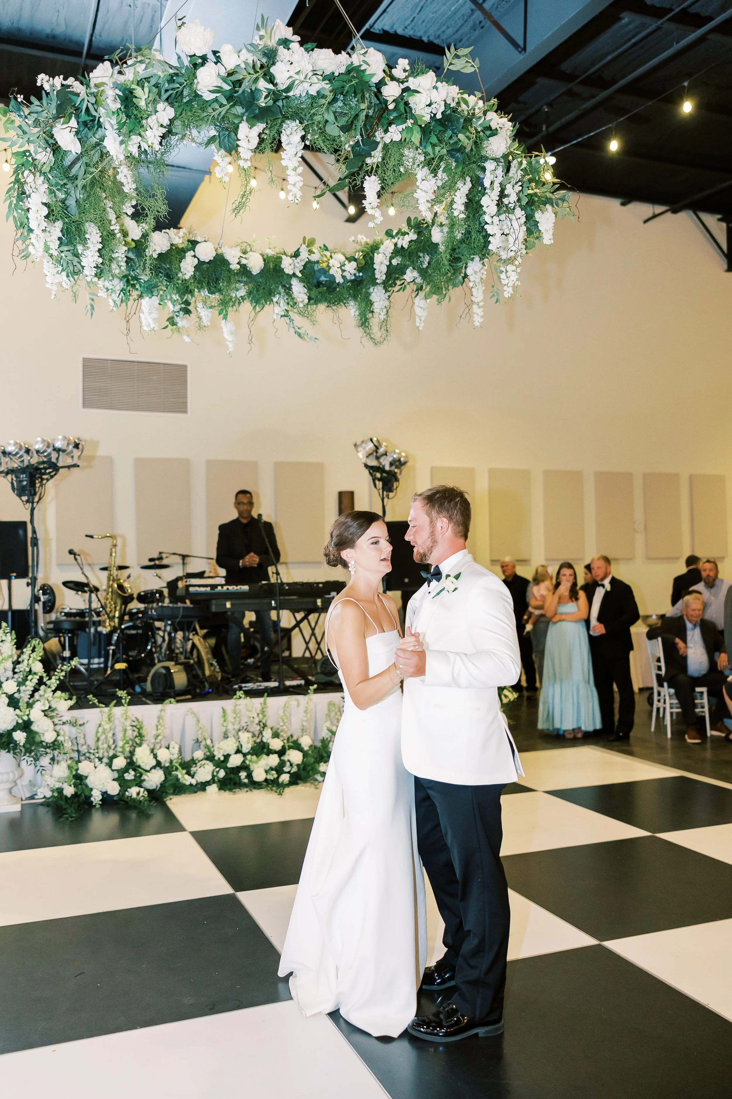 A bride and groom dancing at their wedding reception. The bride is wearing a white gown, and the groom is in a white tuxedo jacket and black trousers. They are holding hands and looking at each other. The background has a band setup with musical inst