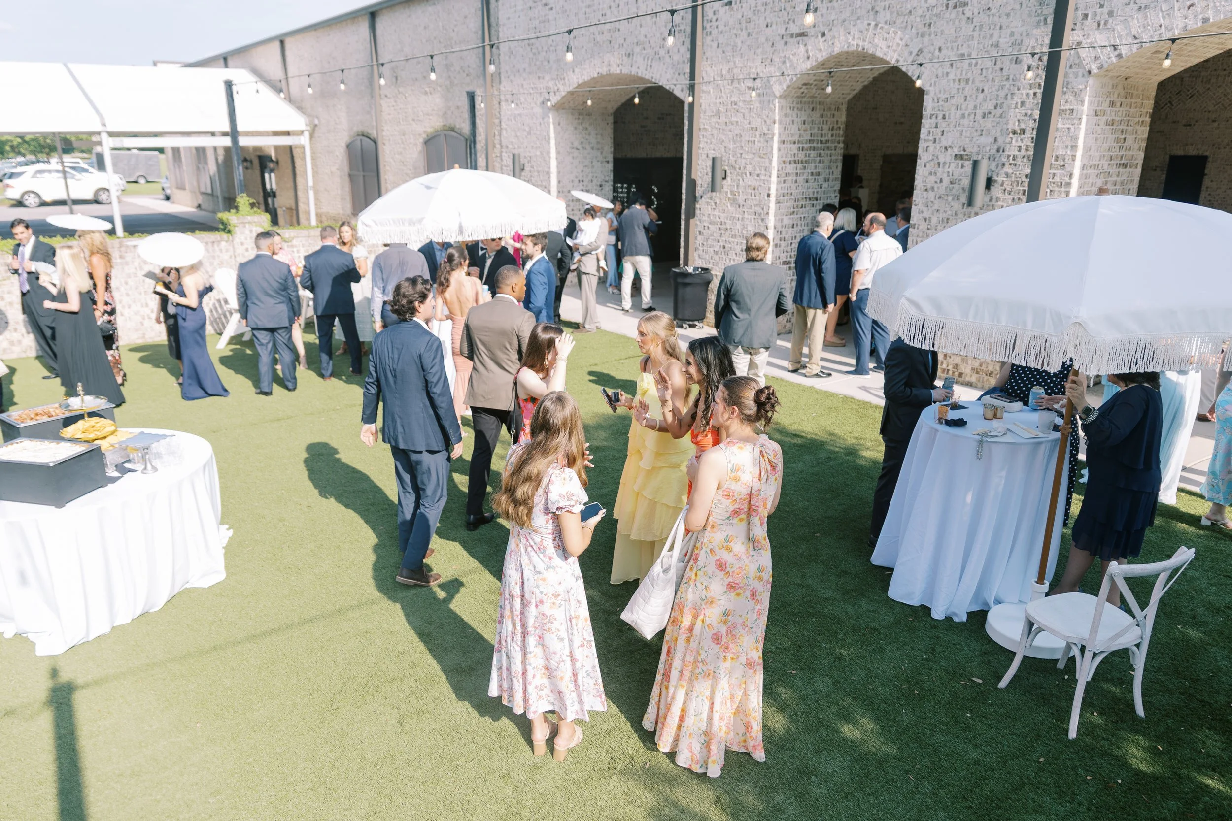 People attending an outdoor wedding reception, standing on grass, some under white umbrellas, dressed in formal and semi-formal attire, with a building in the background.