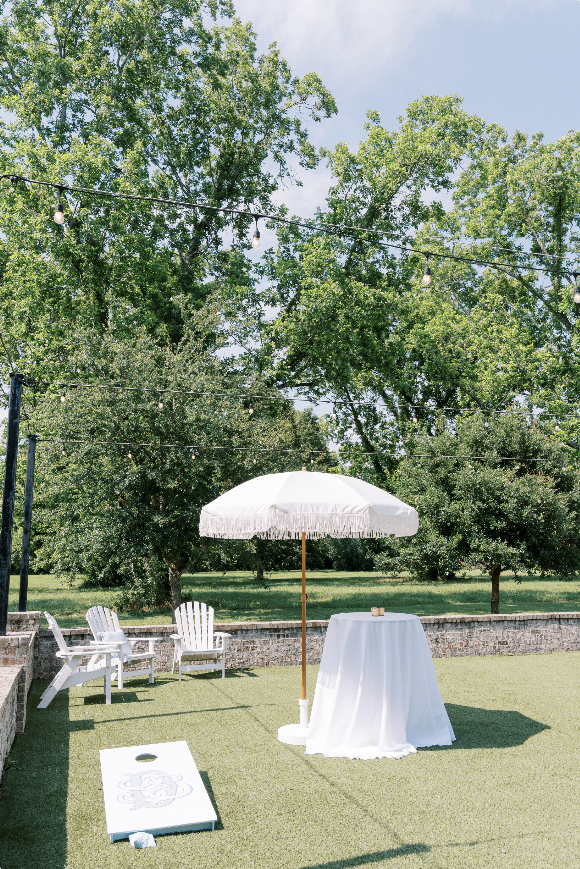 Outdoor patio setup with a white table covered by a cloth, a large white umbrella, two white Adirondack chairs, string lights overhead, and a cornhole game on the grass.