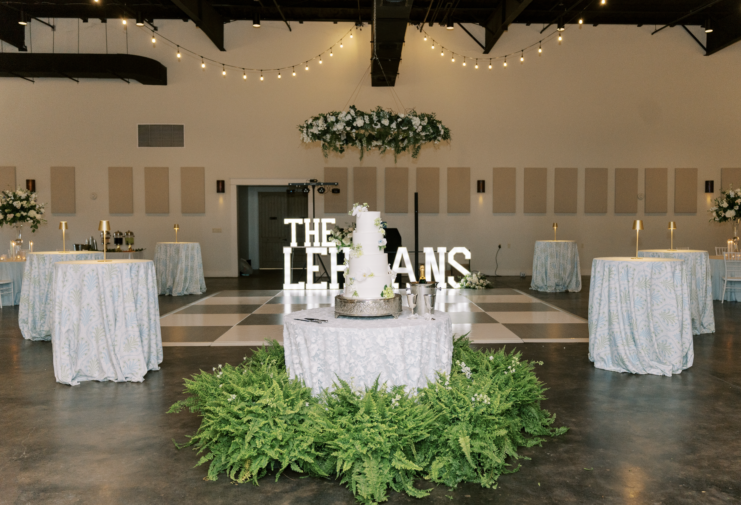 Wedding reception hall with a central wedding cake on a round table surrounded by green ferns, string lights overhead, and tables with floral arrangements. A decorative sign in the background reads "THE LEHANS."