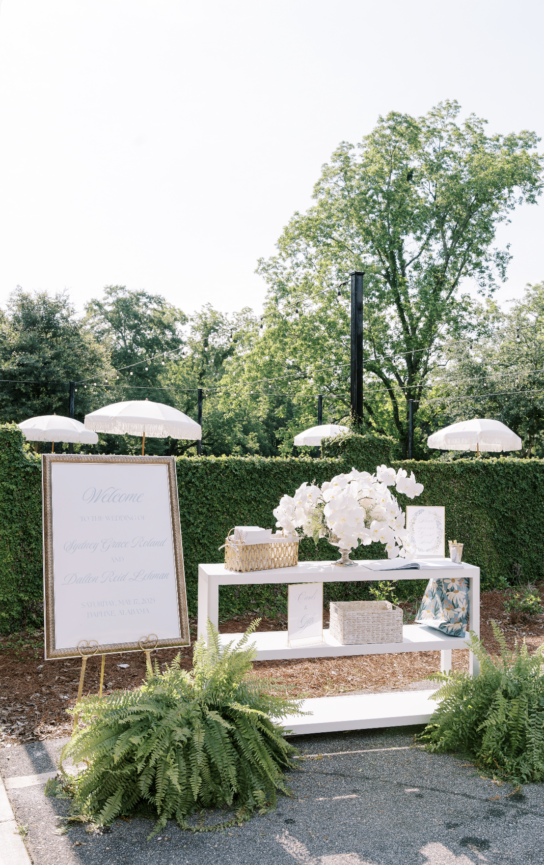 Wedding welcome table outdoors with large white floral arrangement, framed welcome sign, smaller signs, and white umbrellas in the background on a sunny day with trees and greenery.